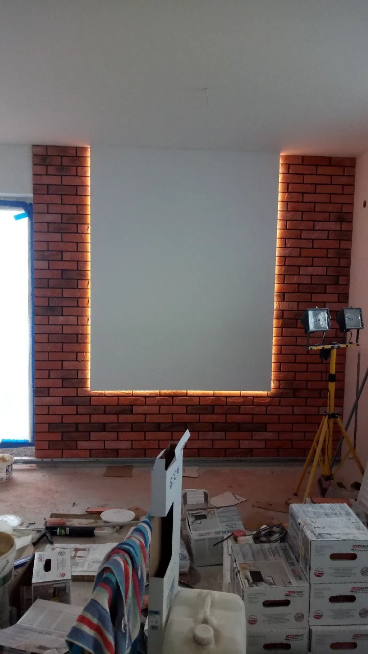 Ściana z cegły klinkierowej z podświetleniem LED tworzącym ramę wokół pomalowanej na biało powierzchni. Na podłodze widoczne materiały budowlane i narzędzia, lampy halogenowe na statywie oświetlają...