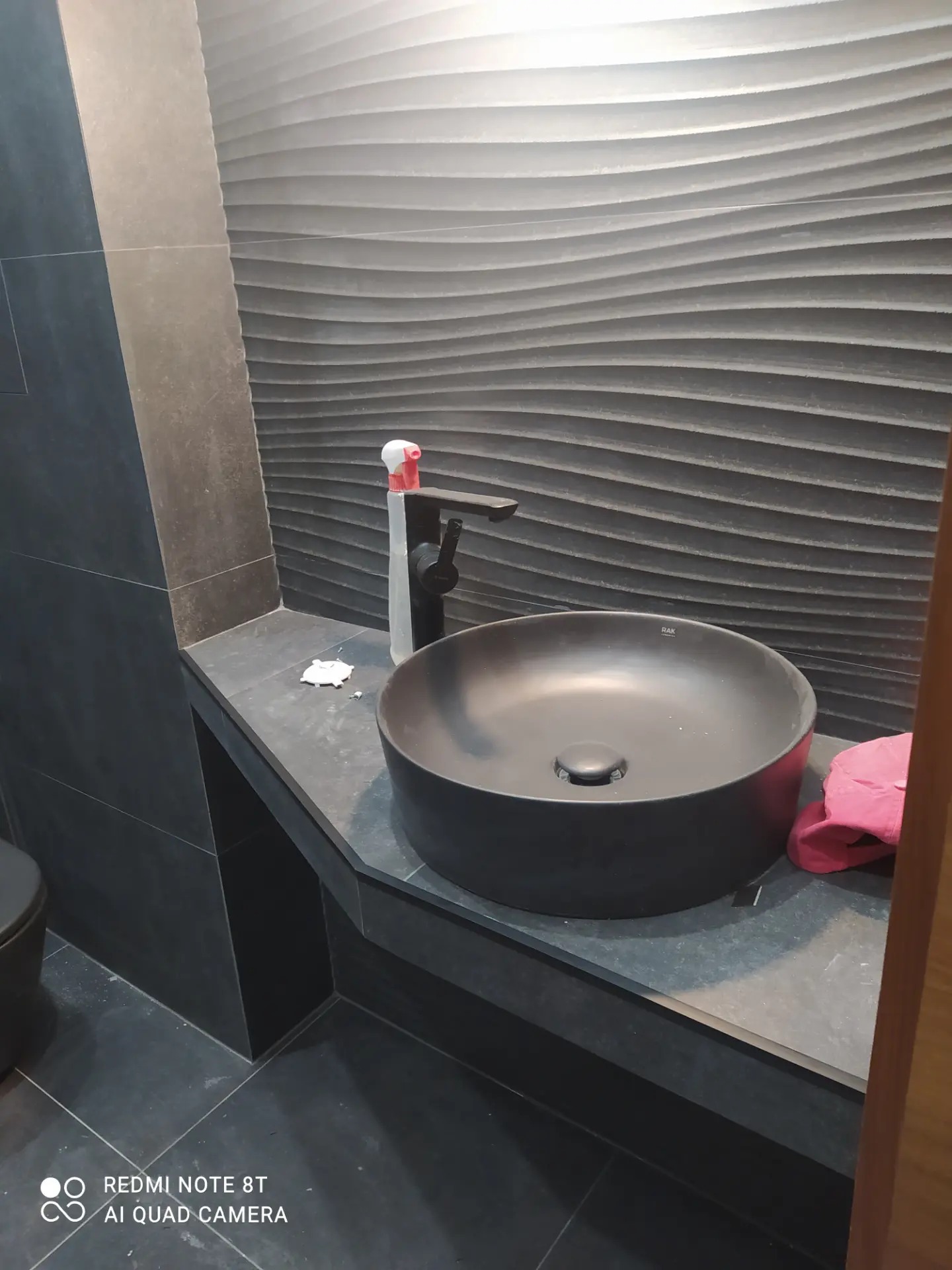 Nowoczesna czarna umywalka nablatowa z czarną armaturą na tle ściany z dekoracyjnymi panelami 3D w odcieniach szarości, widoczne akcesoria łazienkowe i fragment toalety.