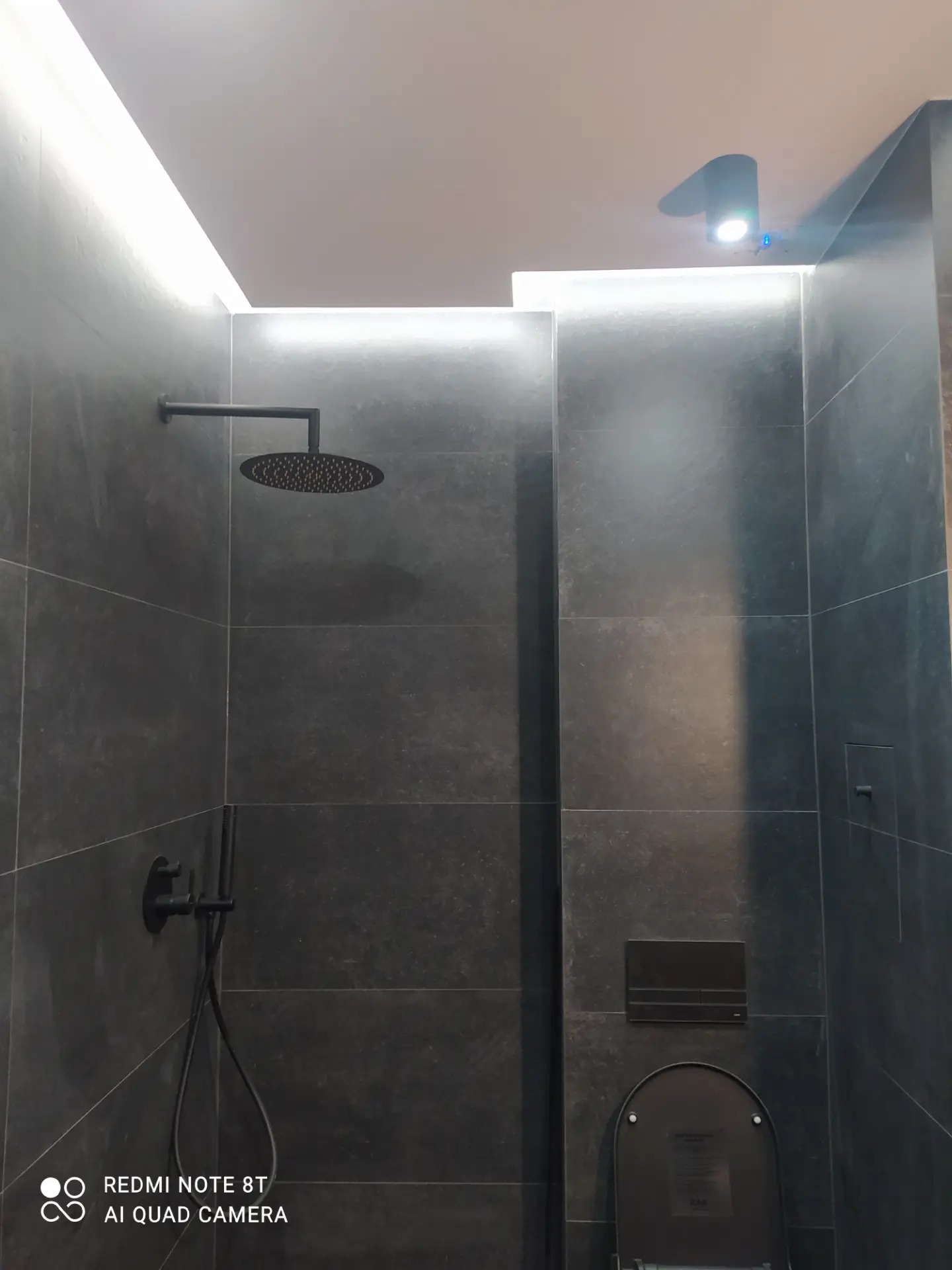 Nowoczesna łazienka z czarnymi płytkami, deszczownicą i podświetleniem LED w suficie.