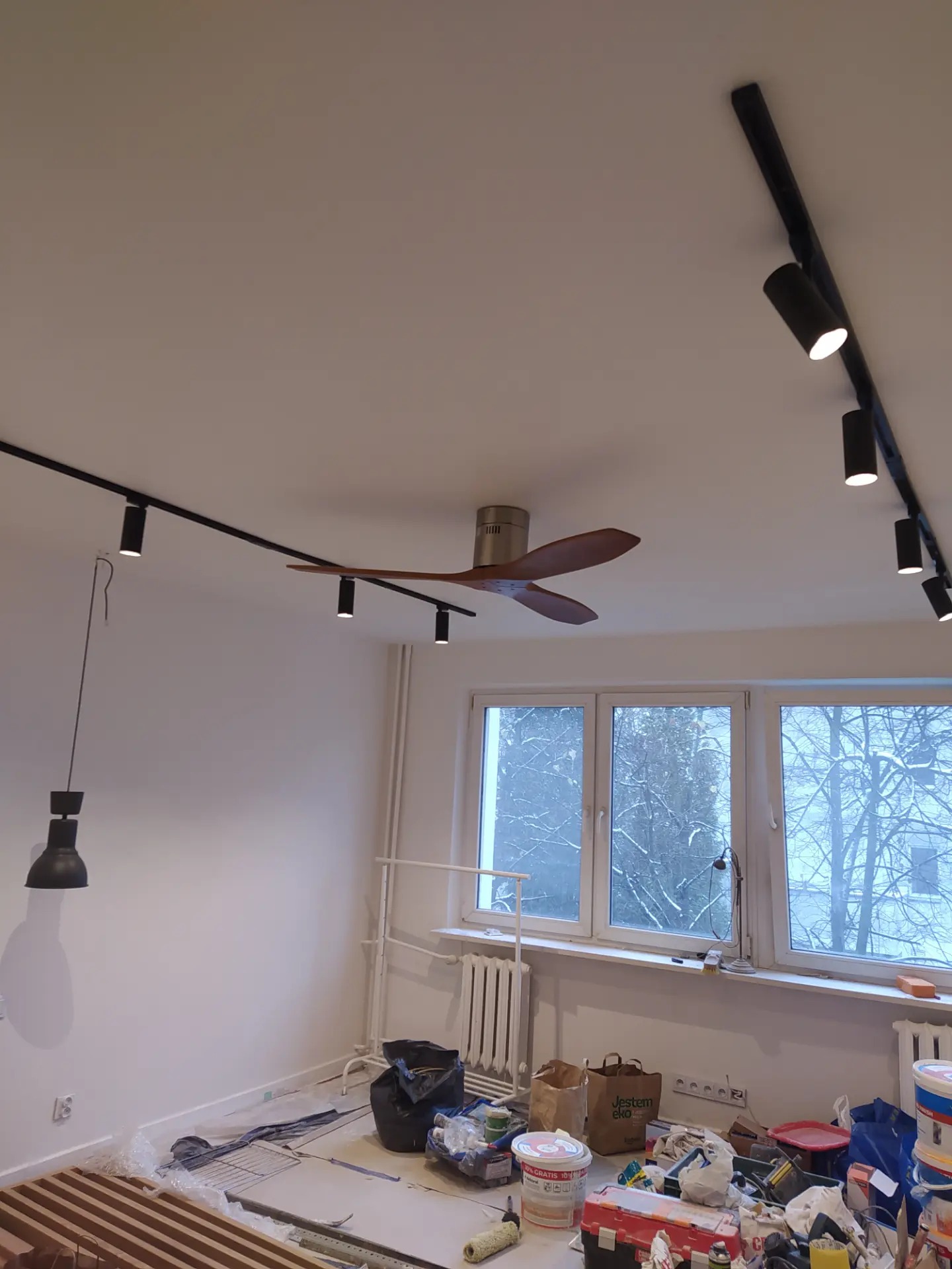 Wnętrze pokoju w trakcie remontu z widocznymi narzędziami malarskimi, listwami drewnianymi na podłodze, lampą wiszącą i szynoprzewodami z reflektorami na suficie oraz wentylatorem sufitowym.