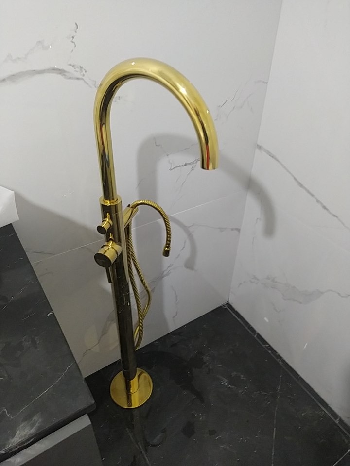 Bateria wannowa wolnostojąca w kolorze złotym, na tle marmurowej ściany i czarnej posadzki z efektem marmuru.