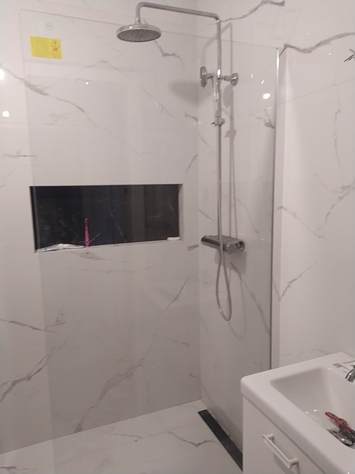 Nowoczesna kabina prysznicowa z deszczownicą, wykończona białymi płytkami imitującymi marmur, wnęka z ciemnym tłem, widoczna umywalka z baterią i narzędzia