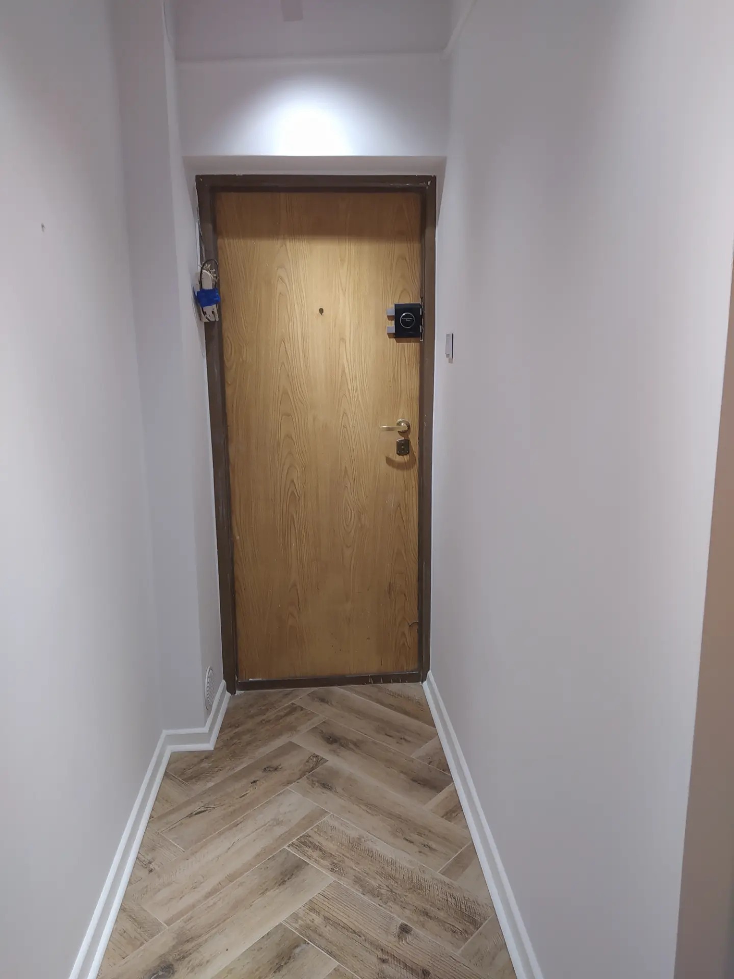 Korytarz po remoncie z drewnopodobną podłogą ułożoną w jodełkę, świeżo pomalowane ściany na jasny kolor, nowe listwy przypodłogowe i drzwi wejściowe z ciemną futryną.