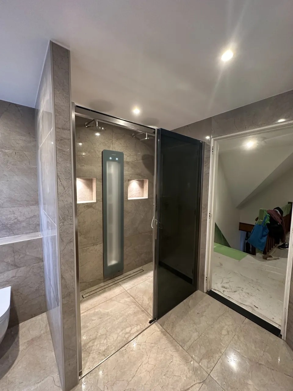 Nowoczesna kabina prysznicowa z szarymi płytkami imitującymi marmur, z wbudowanymi niszami i panelem prysznicowym z podświetleniem, widok na otwarte drzwi do innego pomieszczenia.