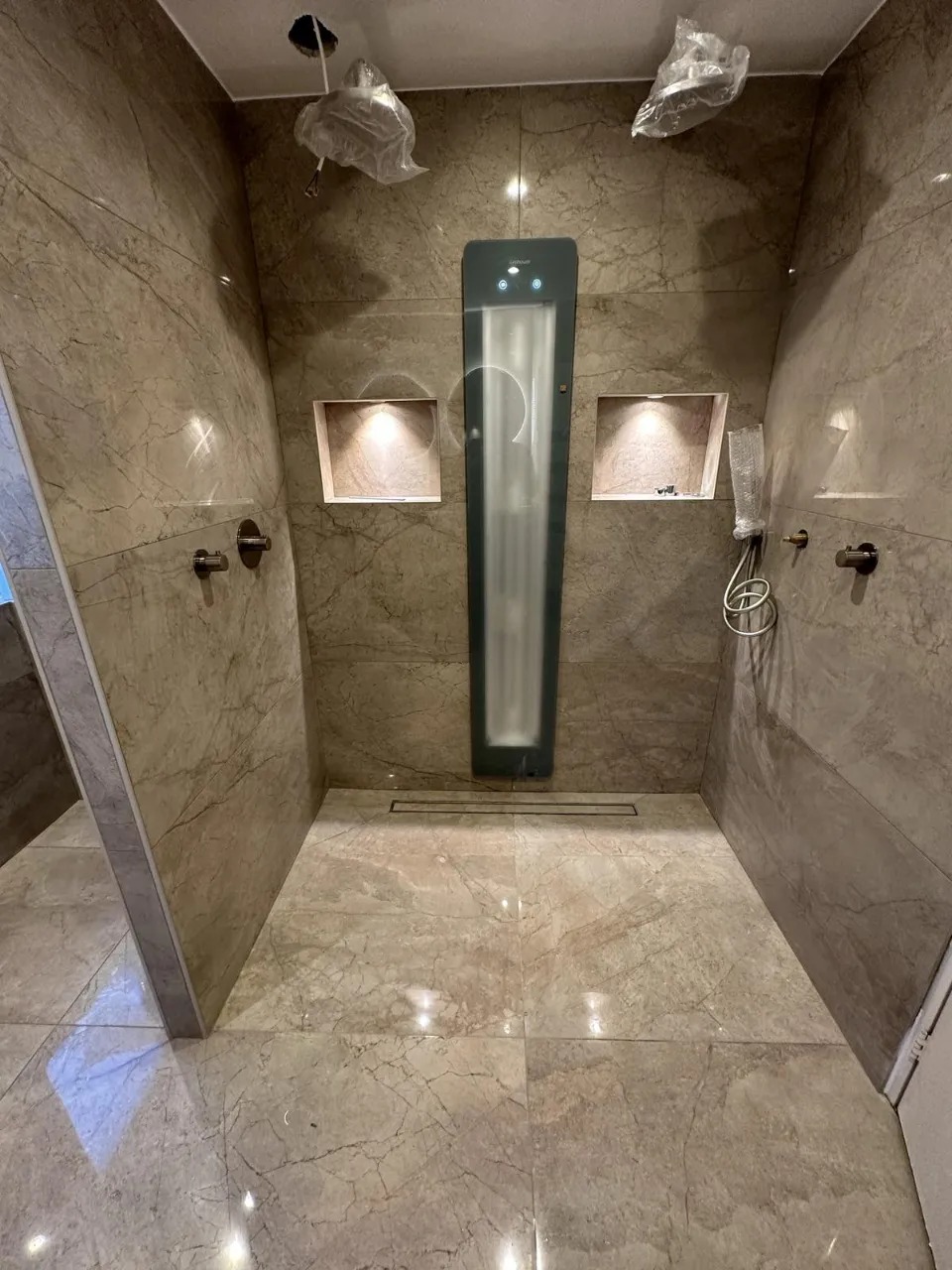 Wykończona kabina prysznicowa walk-in z marmurowymi płytkami w odcieniach beżu i brązu, oświetlone nisze w ścianie, deszczownice w folii ochronnej, odpływ liniowy.