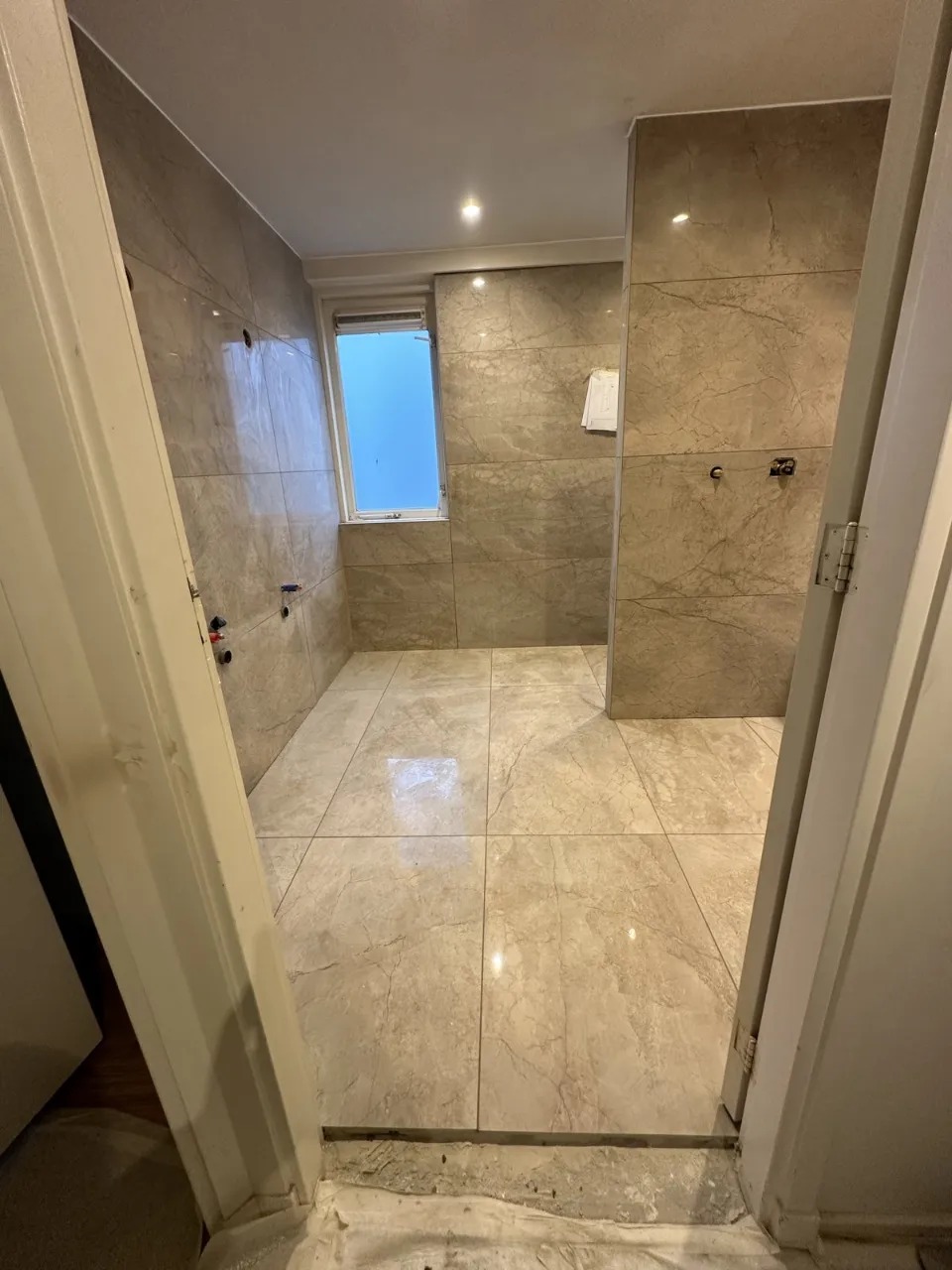 Łazienka w trakcie remontu: położone płytki podłogowe i ścienne o marmurkowym wzorze w beżowej tonacji, widoczne przygotowania do montażu armatury, okno z widokiem na błękitne niebo.