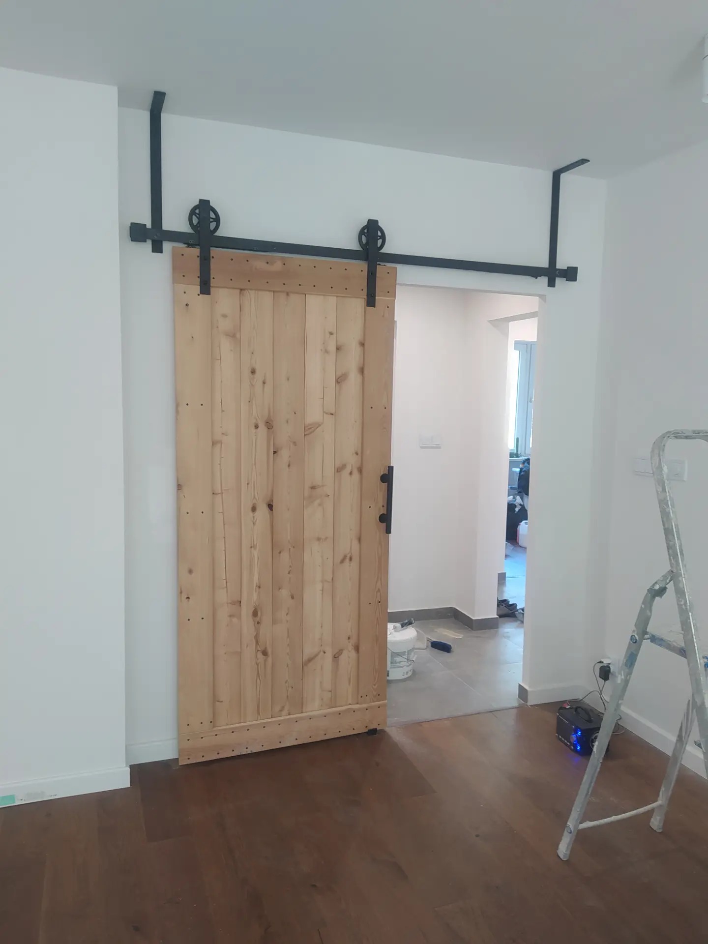 Drewniane drzwi przesuwne w stylu rustykalnym z czarnym systemem mocowania na szynie, widoczne wnętrze mieszkania z podłogą i drabina malarska.