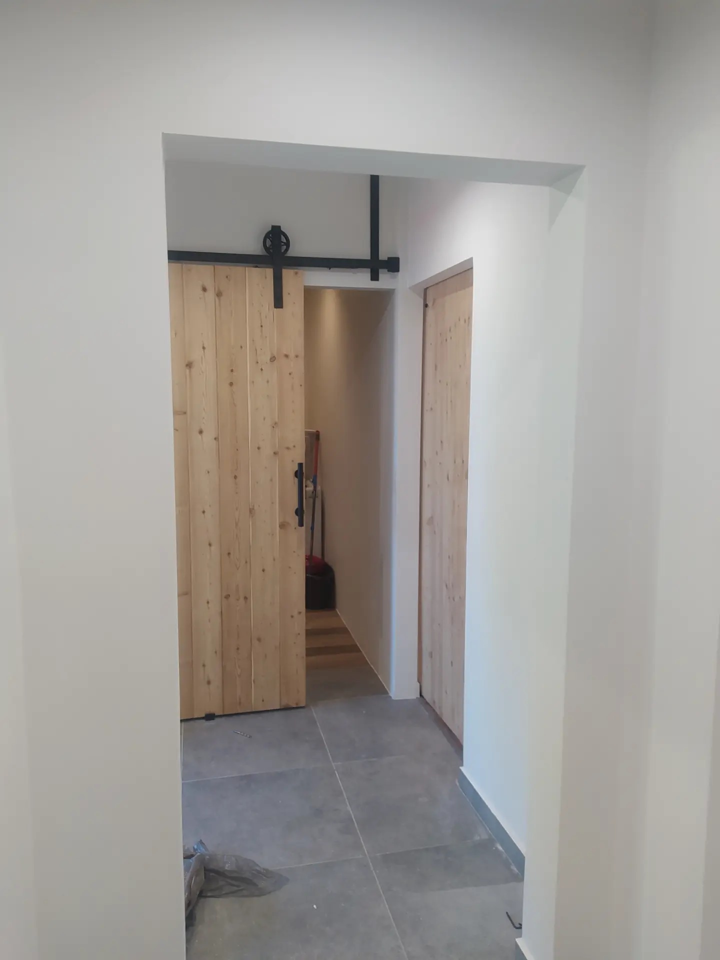 Zamontowane drewniane drzwi przesuwne w stylu rustykalnym z czarnym systemem prowadnic w mieszkaniu w Warszawie. Widoczny fragment podłogi z szarych płytek.