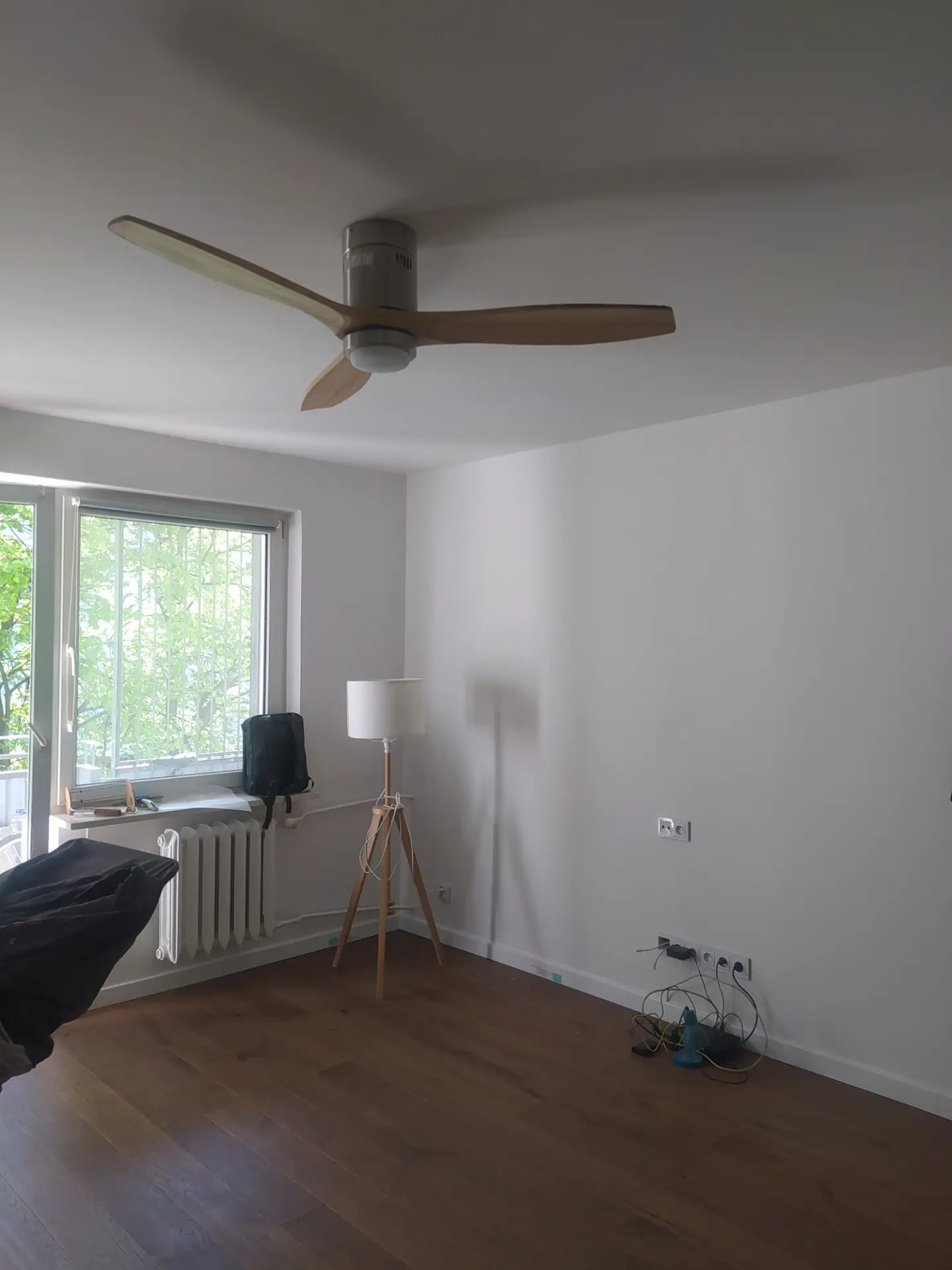 Wnętrze pokoju po remoncie z drewnianą podłogą, białymi ścianami, lampą podłogową na trójnogu, wentylatorem sufitowym z drewnianymi łopatkami i widocznymi gniazdkami elektrycznymi z kablami.