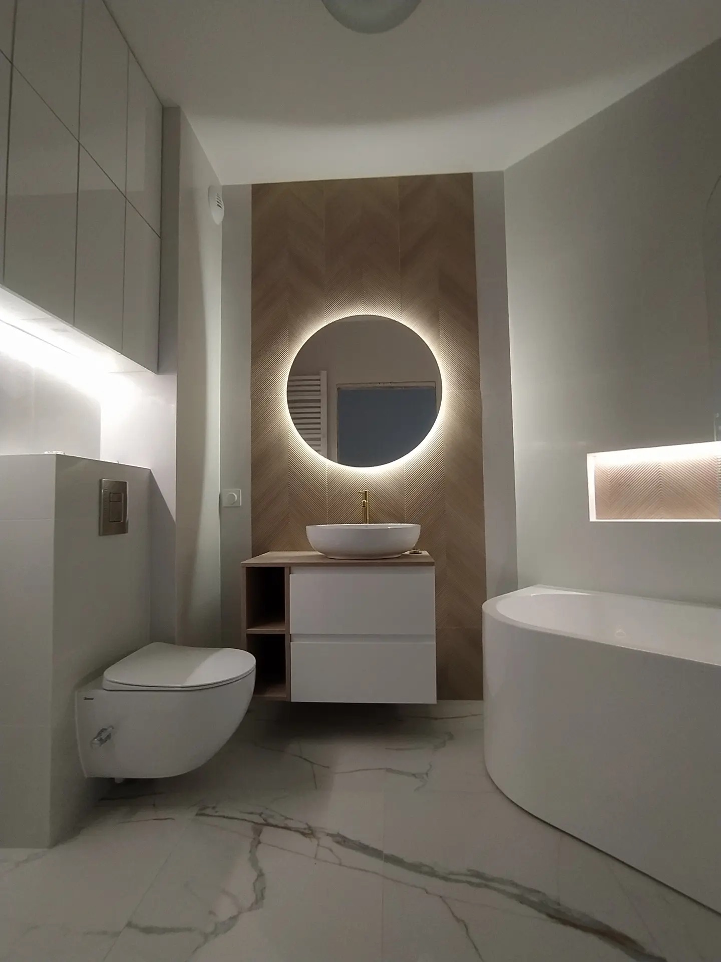 Nowoczesna łazienka z okrągłym lustrem podświetlanym LED, białą umywalką nablatową i armaturą w kolorze złota, płytki imitujące drewno ułożone w jodełkę, oraz podwieszana toaleta i asymetryczna wanna.
