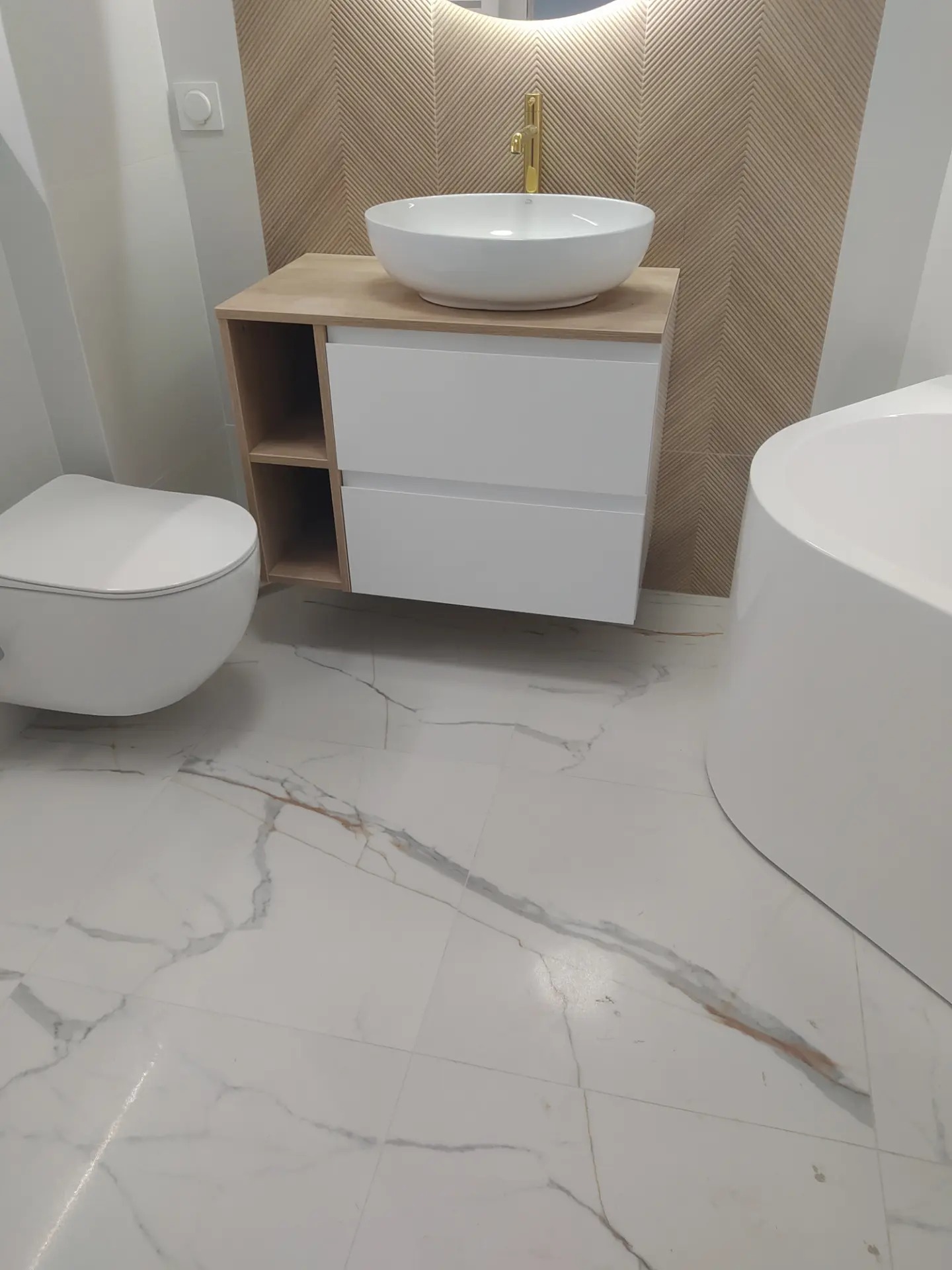 Nowoczesna łazienka z białą ceramiką, umywalką nablatową na szafce z drewnianym blatem, podświetlanym lustrem i płytkami imitującymi drewno ułożonymi w jodełkę na ścianie.