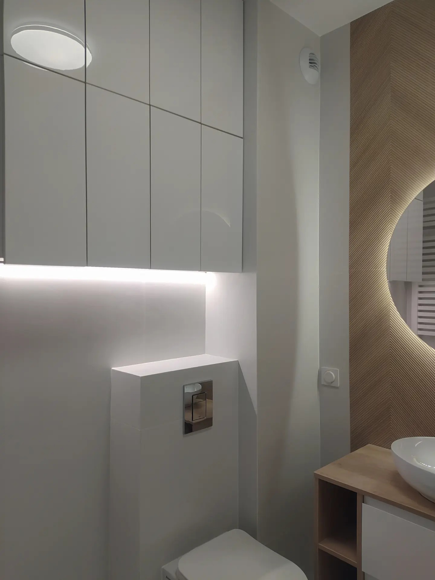 Nowoczesna łazienka z podświetlanymi szafkami nad zabudowaną toaletą, okrągłym lustrem w drewnianej ramie i umywalką nablatową.