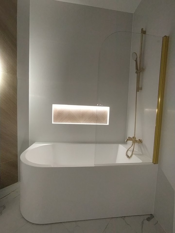 Nowoczesna łazienka z białą wanną z parawanem nawannowym ze złotymi detalami i podświetloną wnęką w ścianie wykończoną wzorem jodełki.