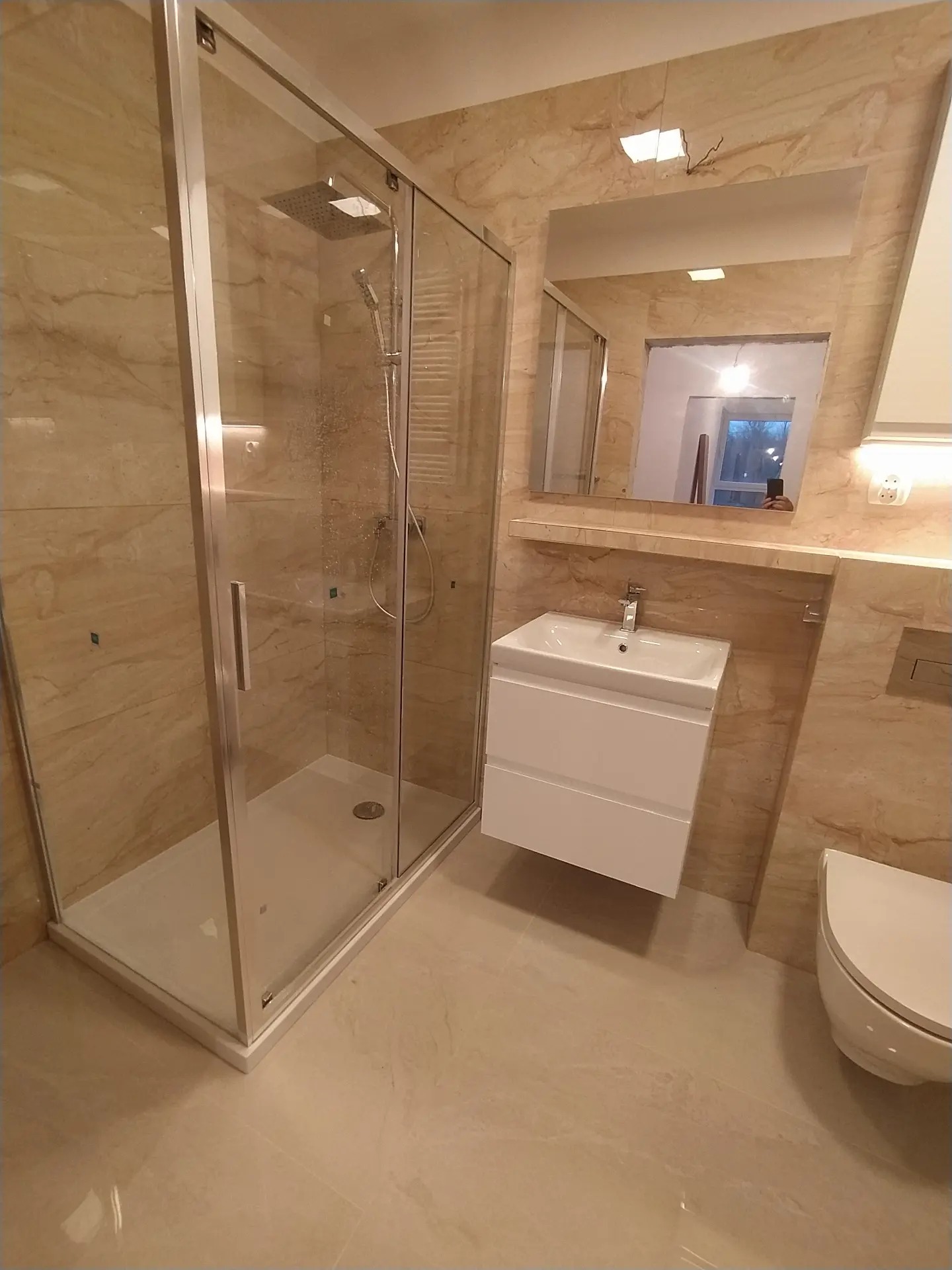 Wykończona łazienka z beżowymi płytkami imitującymi marmur, kabina prysznicowa z chromowanymi profilami, biała umywalka z szafką wiszącą i lustro z oświetleniem górnym.