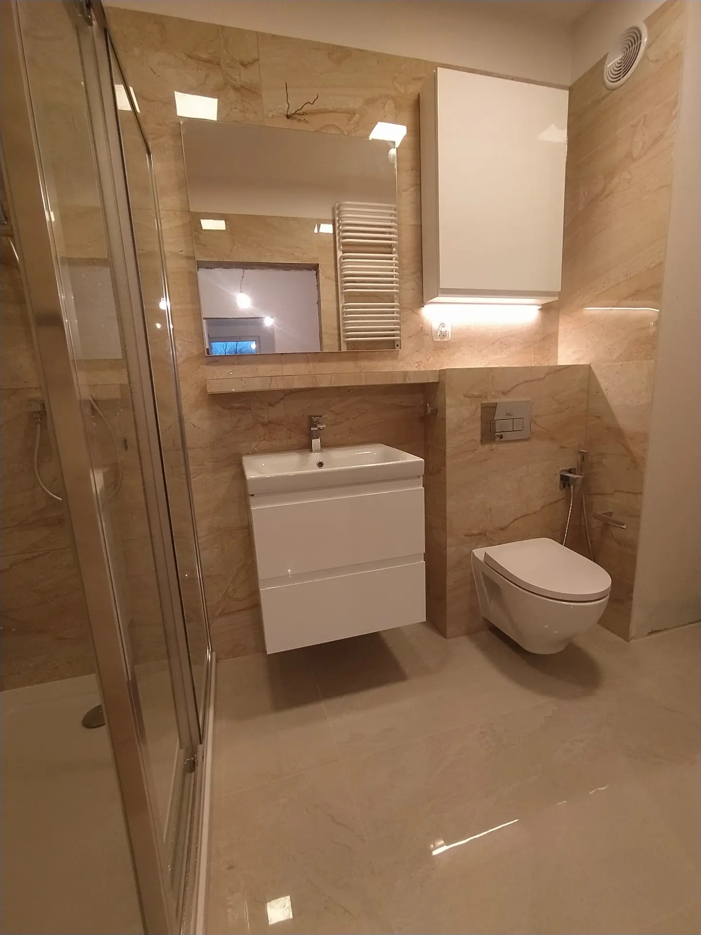 Wykończona łazienka z beżowymi płytkami, wiszącą toaletą i umywalką z szafką, oświetleniem LED pod szafką i lustrem, kabina prysznicowa ze szklanymi drzwiami.