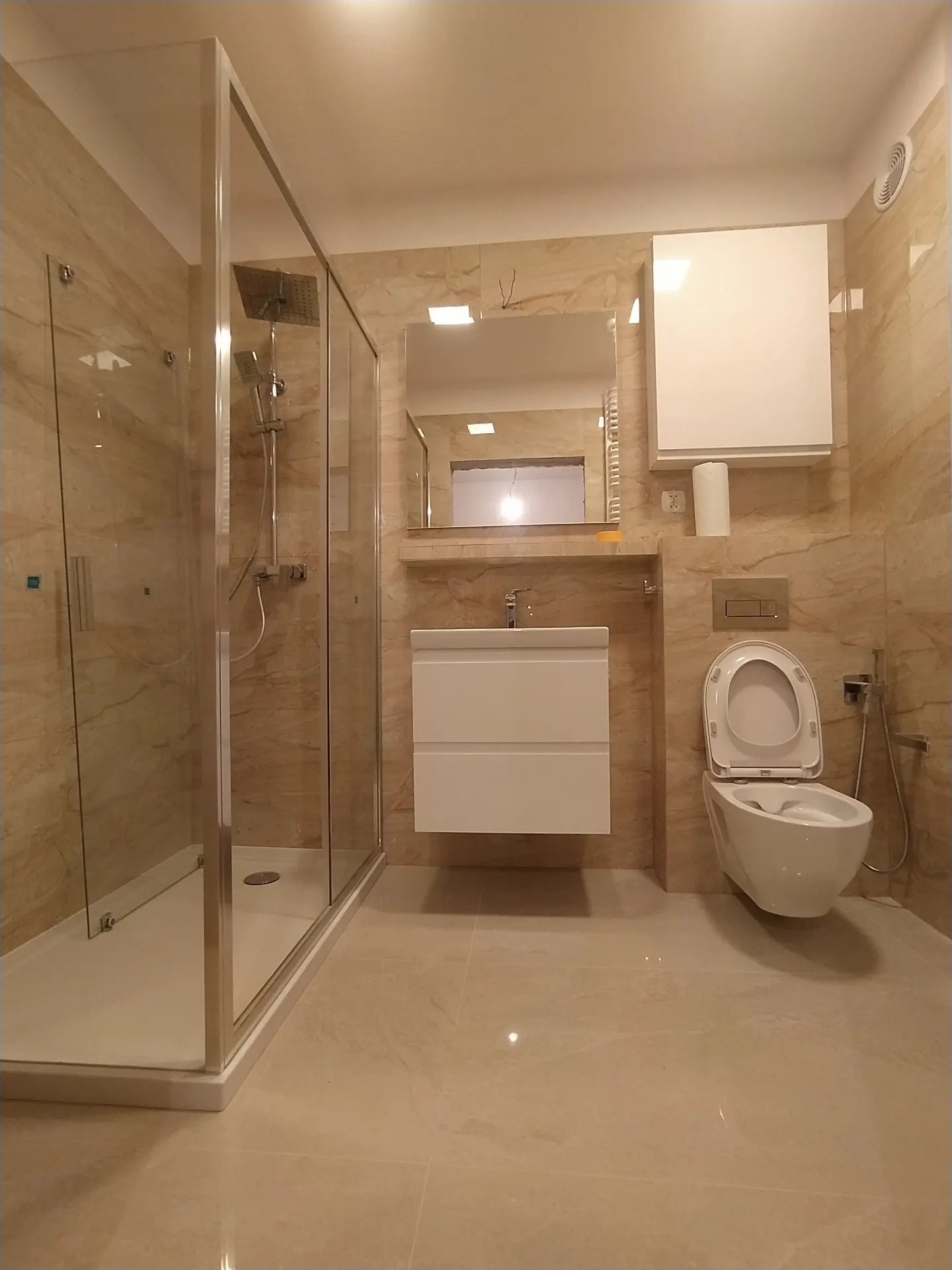 Wykończona łazienka z beżowymi płytkami, kabiną prysznicową typu walk-in, podwieszaną toaletą z bidetką i szafką z umywalką.