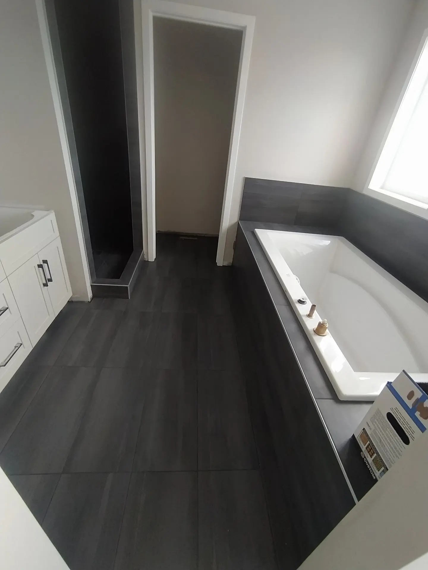 Nowoczesna łazienka z ciemnymi płytkami podłogowymi i prysznicem typu walk-in, biała wanna z baterią w trakcie montażu, oraz biała szafka pod umywalkę.