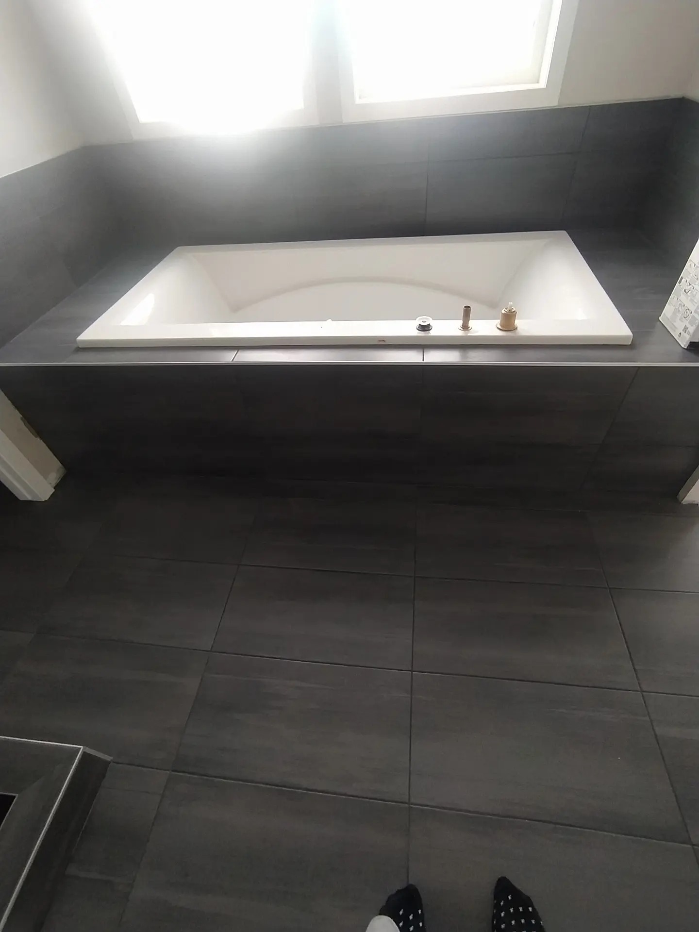 Nowoczesna, wykafelkowana łazienka z wbudowaną białą wanną, widoczne elementy armatury przed montażem i instrukcja obsługi obok wanny, szare płytki na podłodze i ścianach, górne oświetlenie.