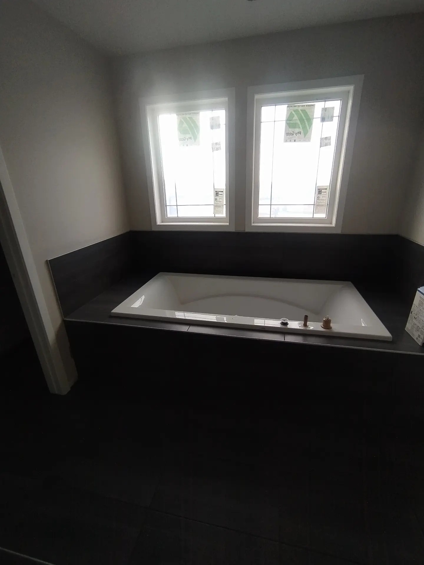 Wanna w obudowie z ciemnych płytek, dwa okna z naklejkami producenta, minimalistyczne wnętrze łazienki w trakcie wykańczania.