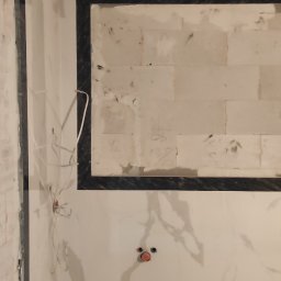 Igor Gonchar - Remont łazienki w trakcie: białe marmurowe płytki na ścianach i podłodze, czarna rama lustra, wystające przewody elektryczne i puszki instalacyjne.