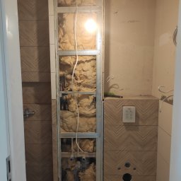 Igor Gonchar - Łazienka w trakcie remontu: widoczna ściana z izolacją z wełny mineralnej w metalowej konstrukcji, obok ściana wykończona płytkami w jodełkę, z zamontowanym panelem sterującym spłuczką i otworami...