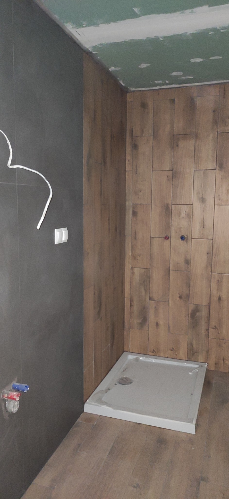 Wnętrze łazienki w trakcie remontu w Radzyminie: widoczne płytki imitujące drewno na ścianie, szare płytki na drugiej ścianie, zamontowany brodzik i wystające kable elektryczne.