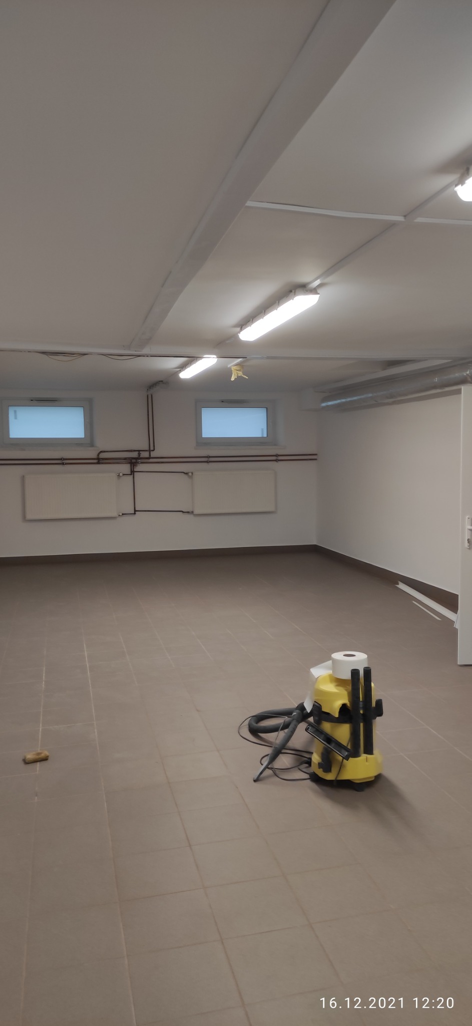 Wnętrze pomieszczenia z ułożoną podłogą z płytek ceramicznych, widoczny odkurzacz przemysłowy Karcher i fragment gąbki na podłodze, grzejniki na ścianie i okna w tle.
