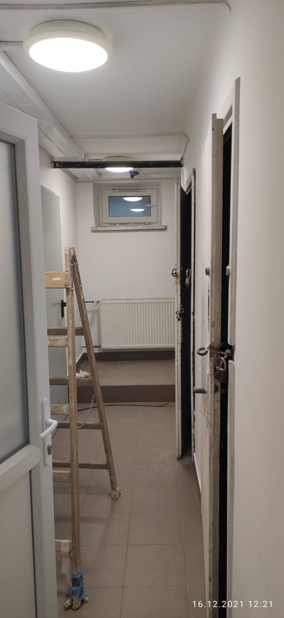 Korytarz w trakcie remontu z drewnianą drabiną opartą o białą futrynę, widoczne otwarte drzwi, grzejnik, okno i lampy sufitowe.