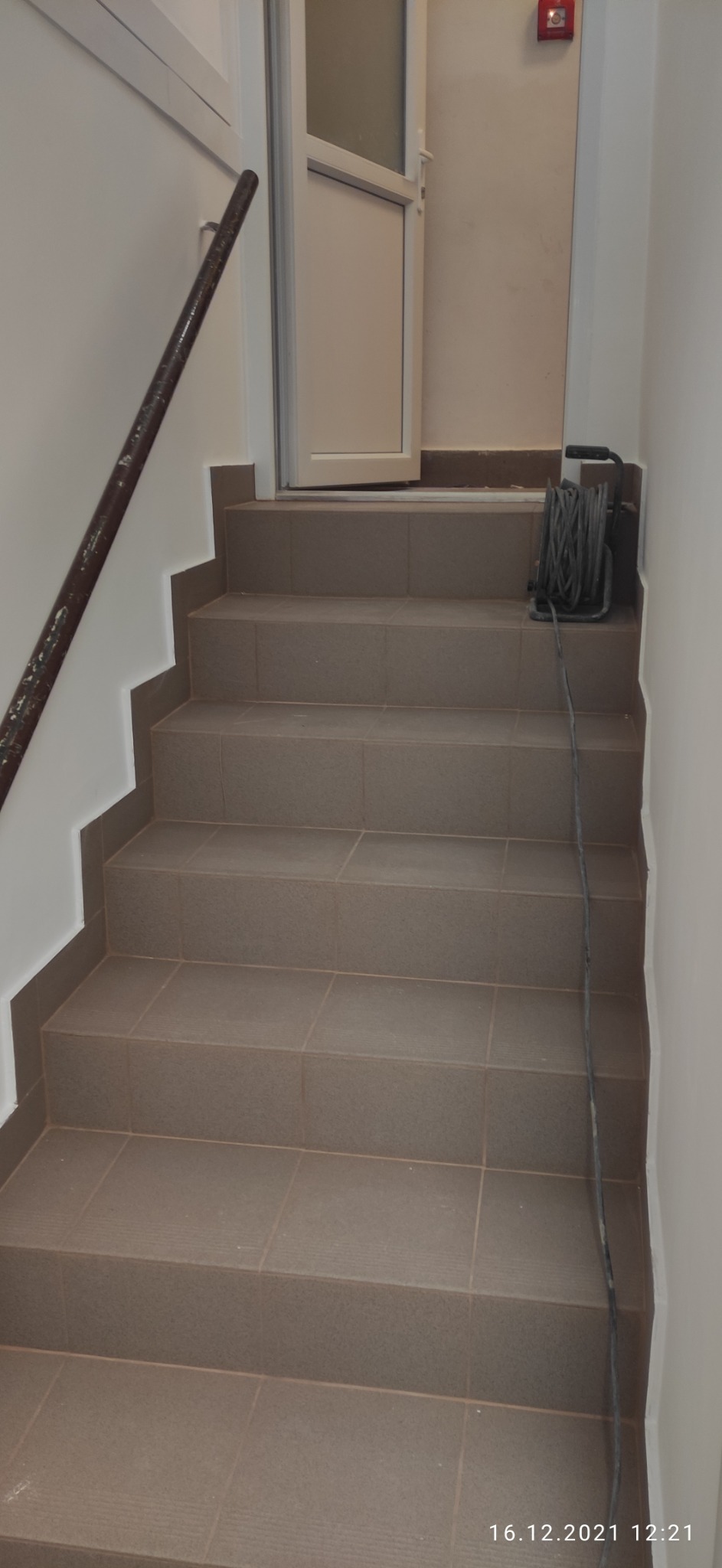 Wykończone schody wewnętrzne wyłożone brązowymi płytkami ceramicznymi, z metalową poręczą po lewej stronie i zwiniętym przewodem elektrycznym na podeście.