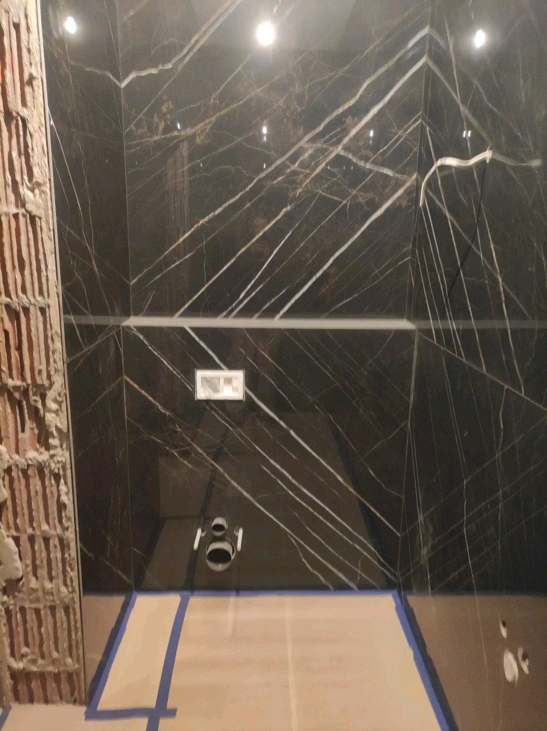 Częściowo wykończona łazienka z czarnymi płytkami imitującymi marmur, widoczne przyłącza do toalety, fragment ściany z odsłoniętą cegłą i podłoga zabezpieczona taśmą malarską.