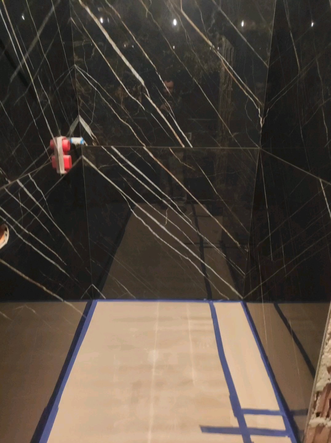 Ściana i podłoga w trakcie układania czarnych płytek z białym żyłkowaniem, zabezpieczone niebieską taśmą malarską, widoczny wystający element instalacji wodnej.