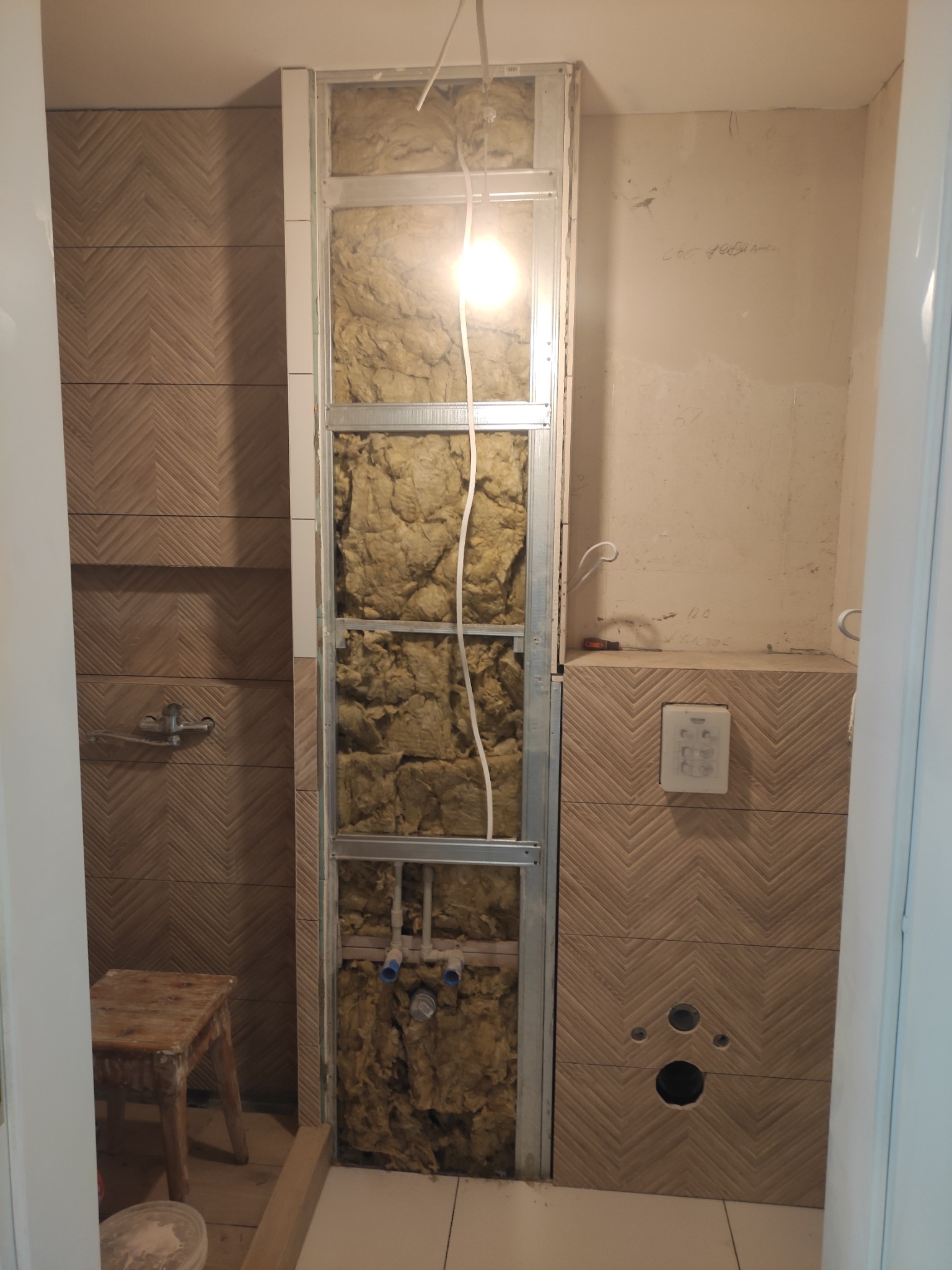 Remont łazienki w trakcie: widoczne płytki imitujące drewno ułożone w jodełkę, metalowa konstrukcja zabudowy z izolacją z wełny mineralnej, instalacja wodna i kanalizacyjna, oraz taboret malarski.