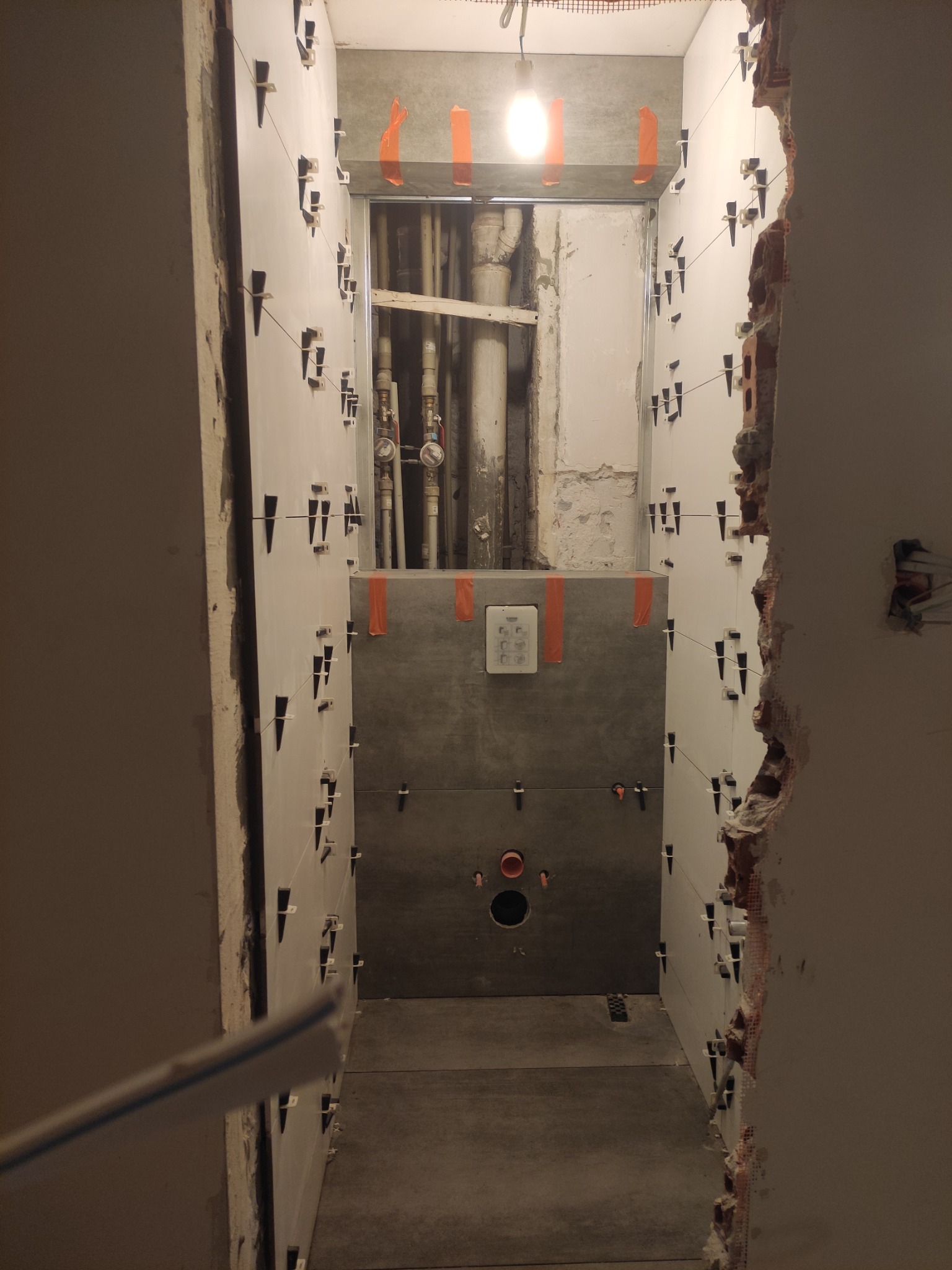 Łazienka w trakcie remontu: białe płytki układane z użyciem klipsów poziomujących, widoczne rury instalacyjne, surowy mur z lewej strony i szare płytki za zabudową WC.