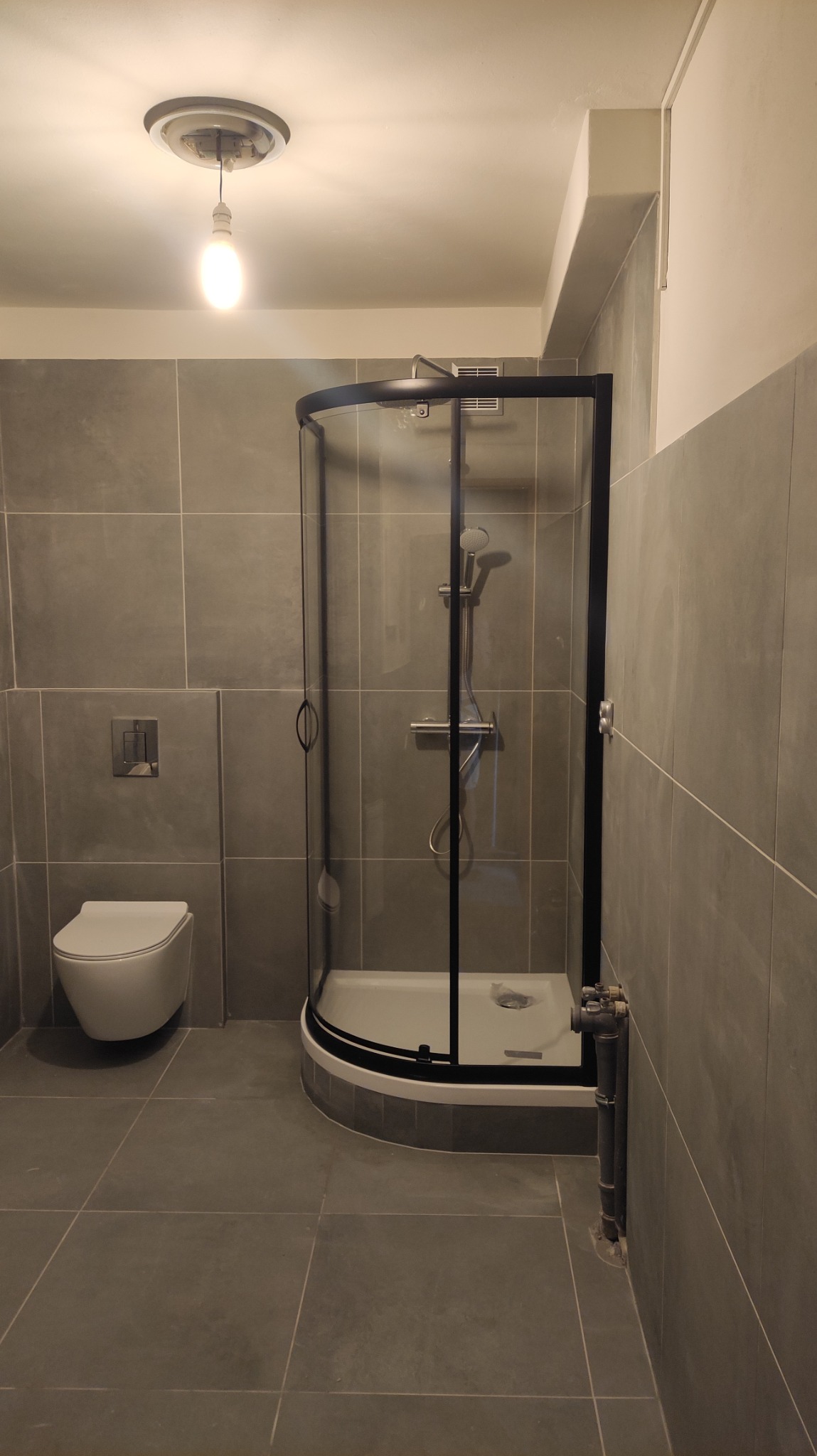 Nowoczesna łazienka z szarymi płytkami, kabiną prysznicową z czarnymi ramami i podwieszaną toaletą.