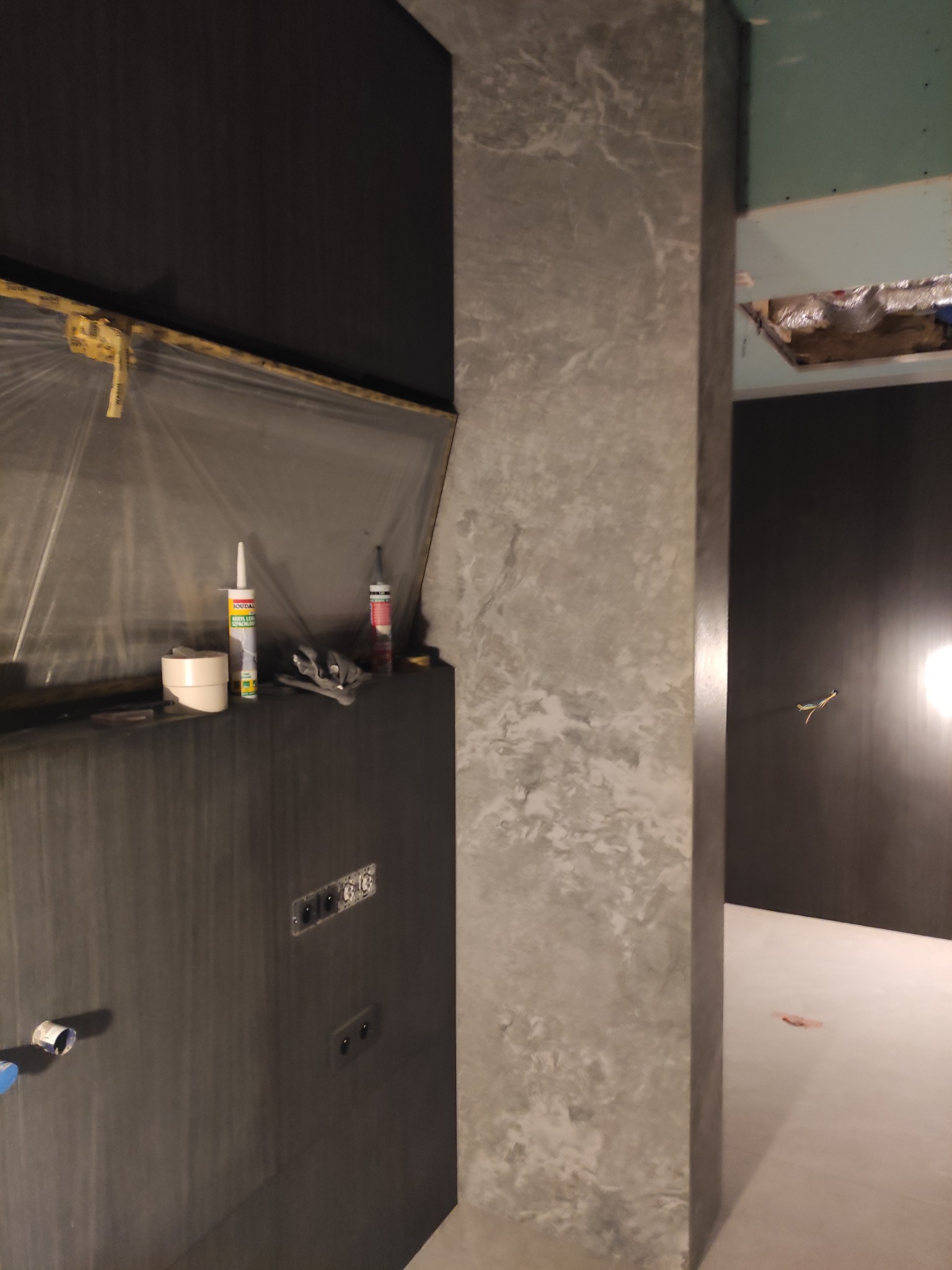 Łazienka w trakcie remontu: widoczna czarna ściana z przygotowanymi otworami na gniazdka, zabezpieczona folią, obok kolumna z szarą, marmurkową strukturą, w tle surowy stan wykończenia.