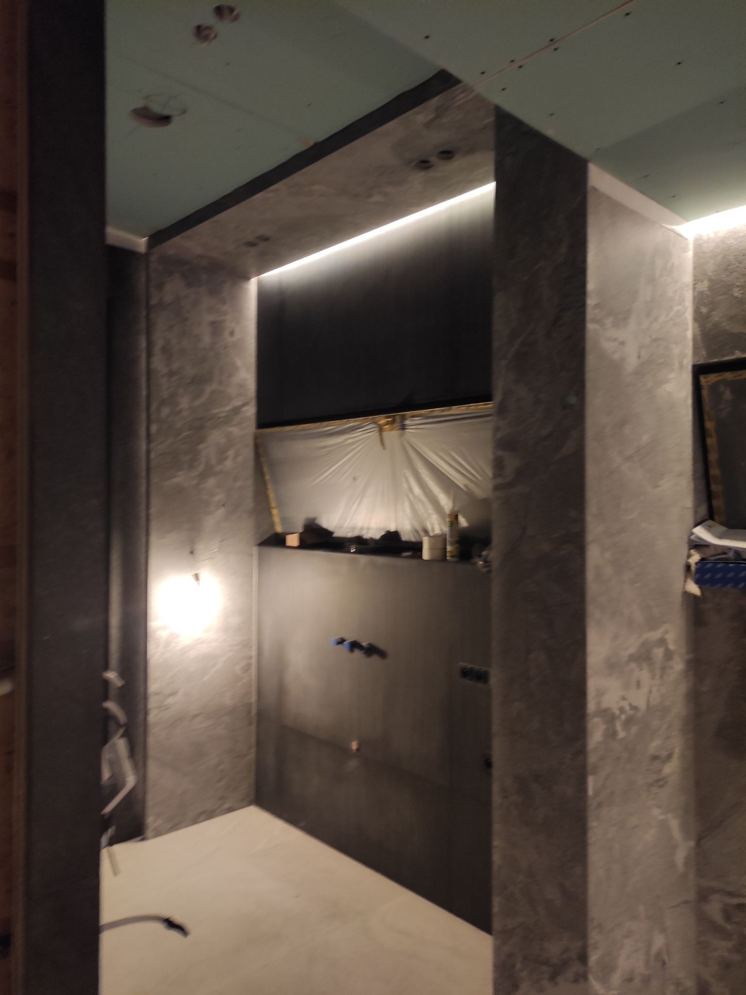 Wnętrze łazienki w trakcie remontu, ściany wykończone w szarym odcieniu z efektem betonu, widoczne przygotowania do montażu oświetlenia LED w niszy, podwieszany sufit w kolorze miętowym, zakryte...