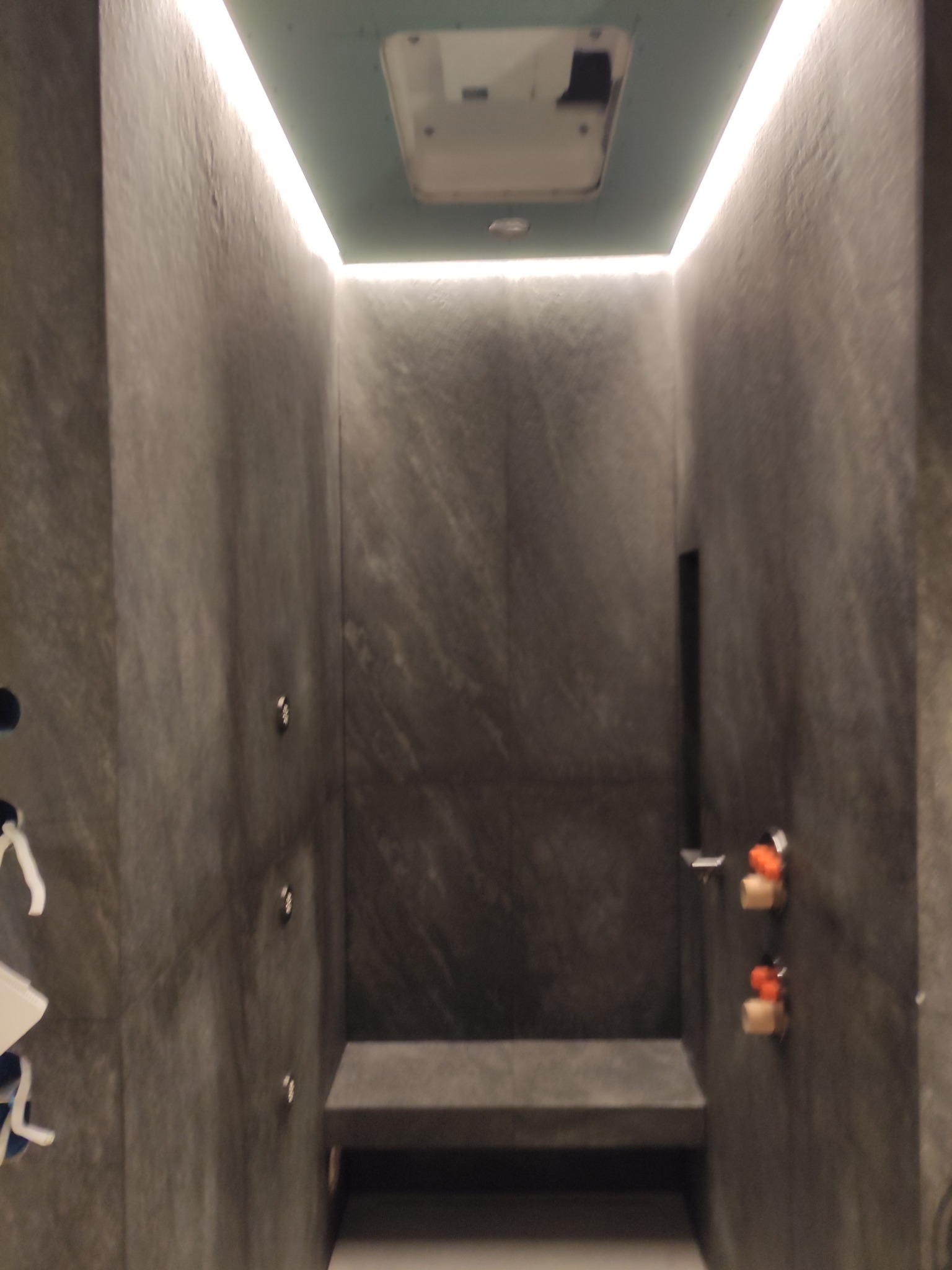 Nowoczesna kabina prysznicowa z ciemnymi, strukturalnymi płytkami, podświetleniem LED wzdłuż sufitu i wnęką z siedziskiem, widoczne elementy instalacji wodnej.