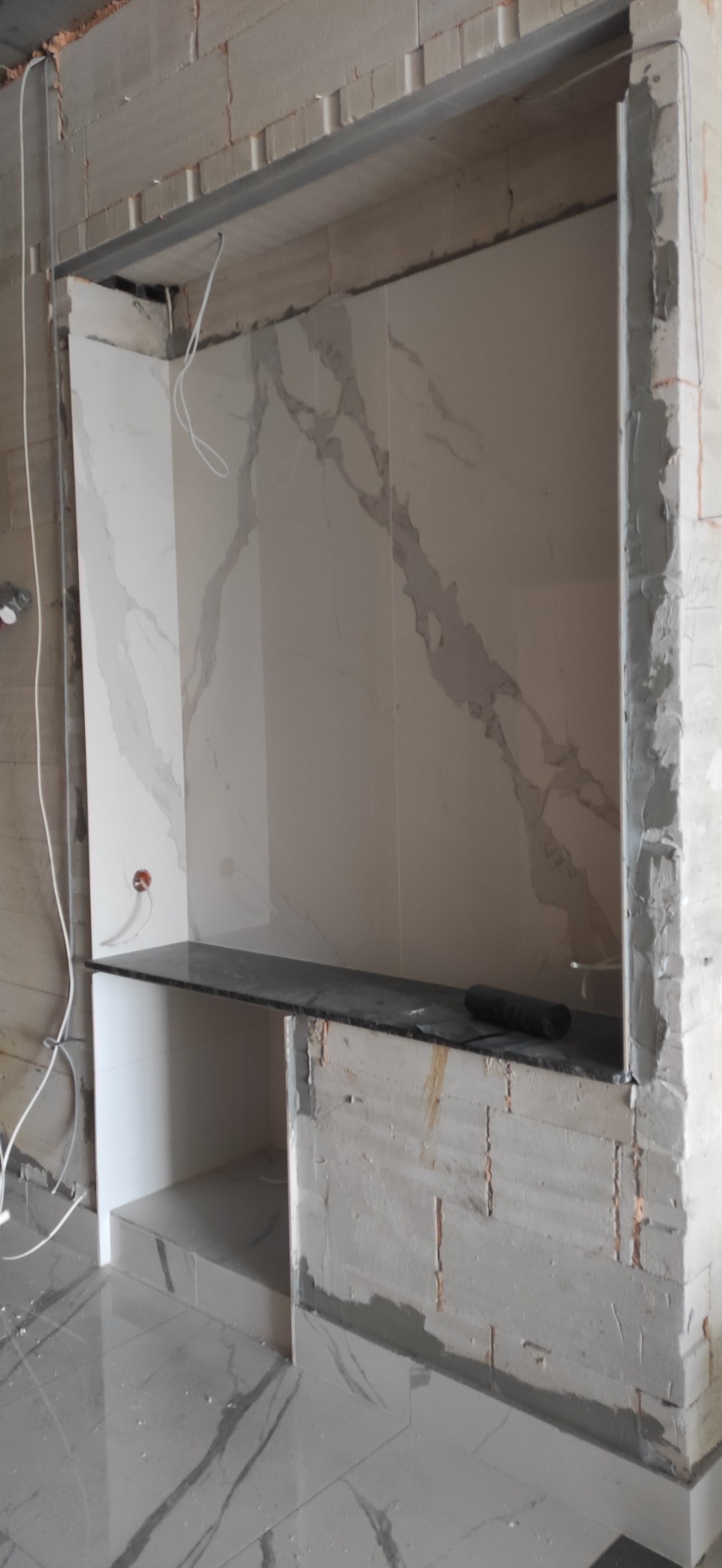 Fragment łazienki w trakcie remontu: ułożone płytki imitujące marmur na ścianie i podłodze, widoczne surowe ściany z betonu komórkowego, półka z ciemnego kamienia, wystające przewody elektryczne.