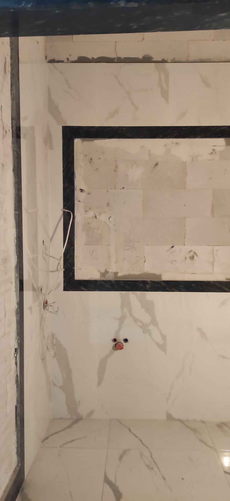 Remont łazienki w trakcie: białe marmurowe płytki na ścianach i podłodze, czarna rama lustra, wystające przewody elektryczne i puszki instalacyjne.