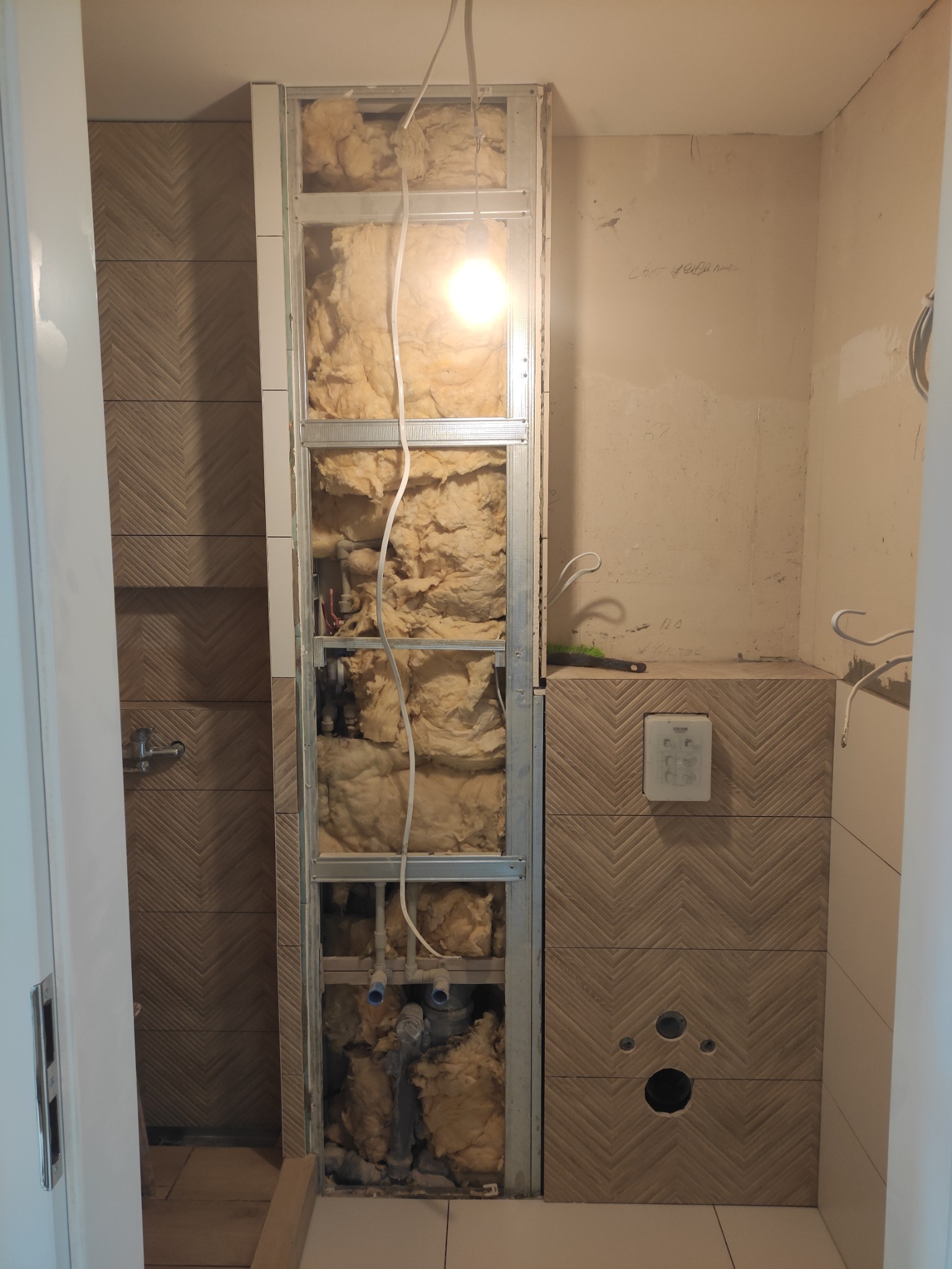 Łazienka w trakcie remontu: widoczna ściana z izolacją z wełny mineralnej w metalowej konstrukcji, obok ściana wykończona płytkami w jodełkę, z zamontowanym panelem sterującym spłuczką i otworami...