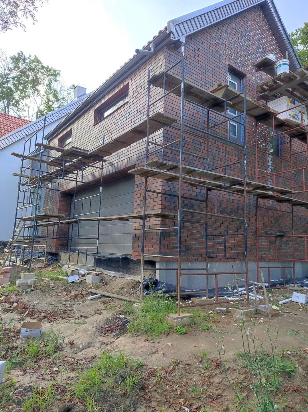 Budynek w trakcie wykańczania elewacji z czerwonej cegły, widoczne rusztowania, częściowo ocieplony garaż, teren wokół budynku w trakcie prac ziemnych.