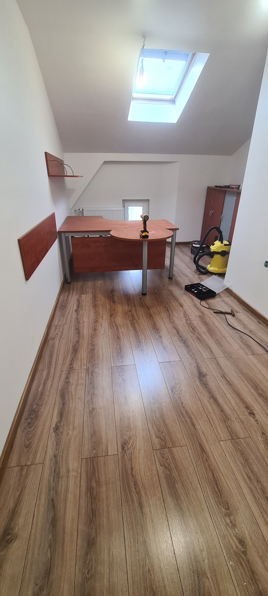 Pokój na poddaszu z drewnianą podłogą, biurkiem z okrągłym blatem i wkrętarką na nim, widoczny odkurzacz przemysłowy i skrzynka z narzędziami, oświetlony przez okno dachowe.