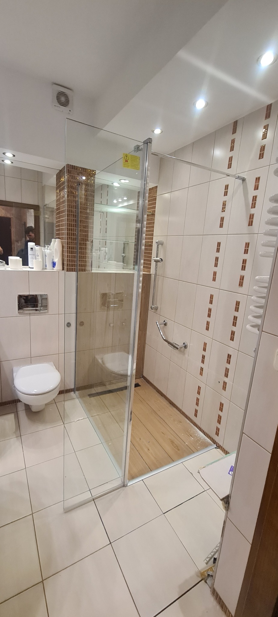 Nowoczesna kabina prysznicowa z transparentnego szkła, zamontowana w łazience z beżowymi płytkami i drewnopodobną podłogą, widoczna toaleta i umywalka z białymi kosmetykami w tle.