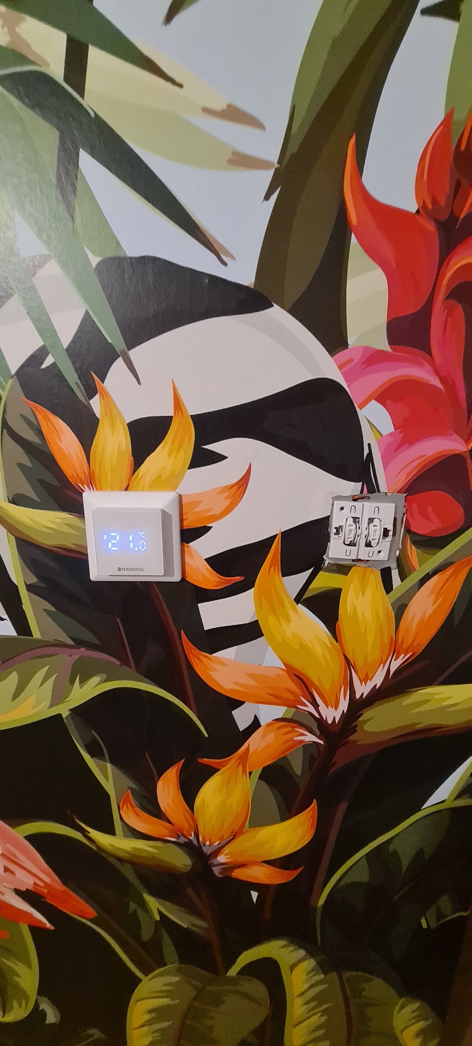 Ściana z tapetą w motywy tropikalne, na której zamontowany jest biały termostat z wyświetlaczem wskazującym 21.8 stopni Celsjusza oraz rozmontowany podwójny włącznik światła z widocznymi przewodami.