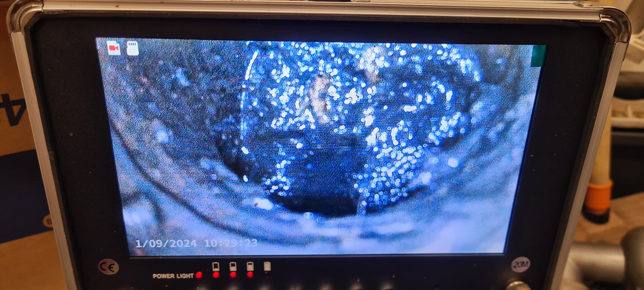 Ekran kamery inspekcyjnej pokazuje wnętrze rury z widocznymi zanieczyszczeniami i wodą, data i godzina na ekranie 1/09/2024 10:29:23, w tle elementy otoczenia miejsca pracy.