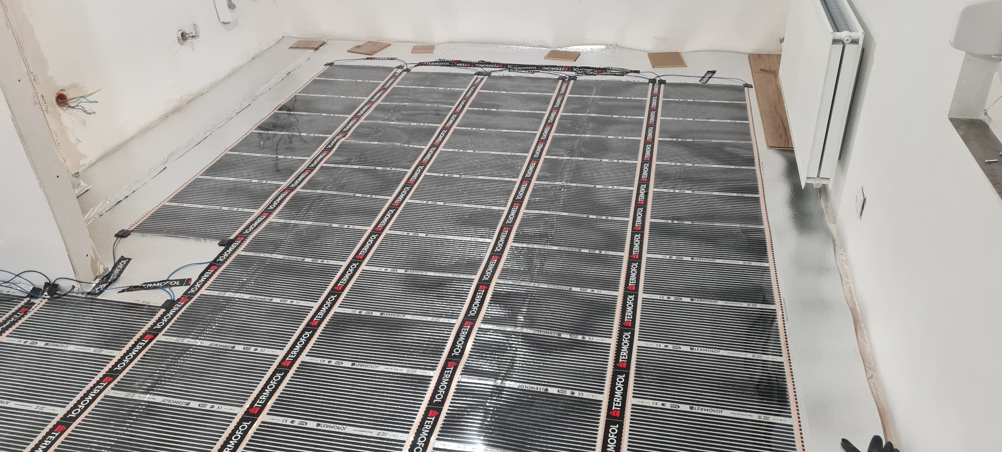 Rozłożona mata grzewcza Termofol na podłodze w surowym stanie, przygotowana do zalania wylewką, widoczne kable i puszka elektryczna w ścianie.