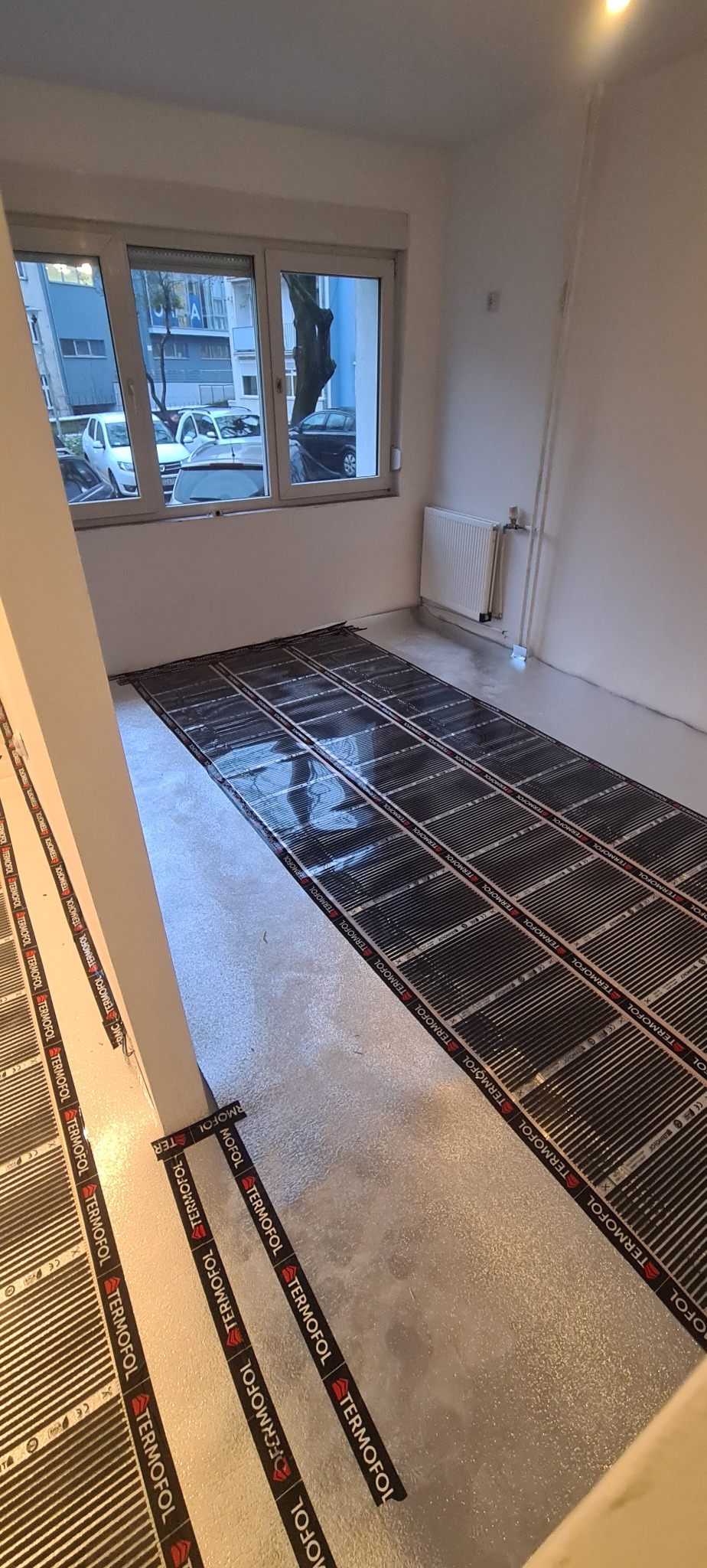 Montaż folii grzewczej Termofol na podłodze w pomieszczeniu przed wylaniem wylewki, widok na okno z widokiem na ulicę i budynki.