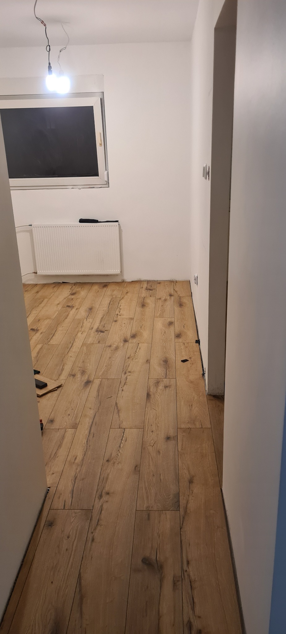 Układanie paneli podłogowych w pokoju z oknem i grzejnikiem, widoczne niedociągnięcia przy ścianie i brak listew przypodłogowych.