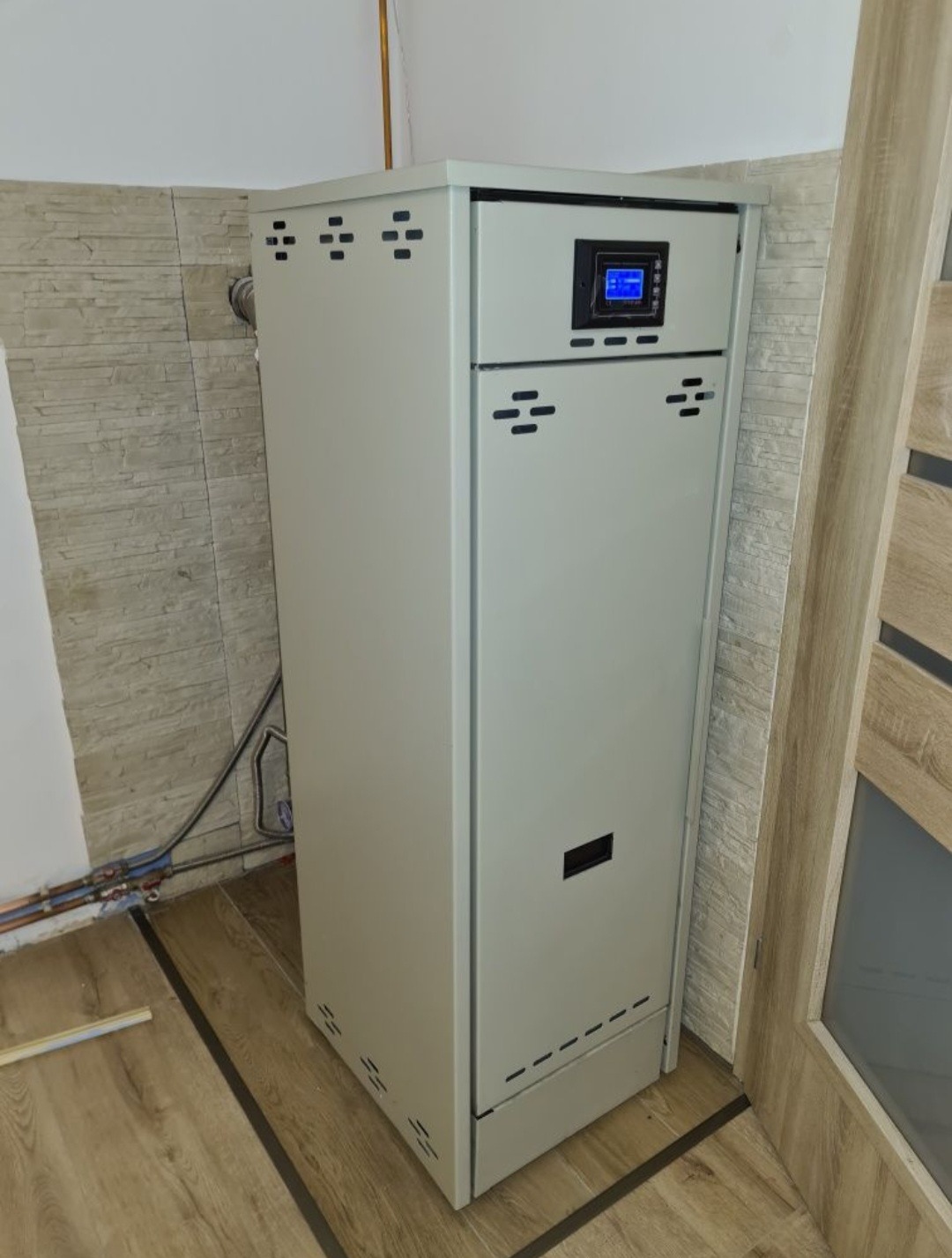 Nowoczesny piec centralnego ogrzewania z elektronicznym panelem sterowania, zainstalowany w pomieszczeniu z drewnopodobną podłogą i częściowo wykończoną ścianą z imitacją kamienia.