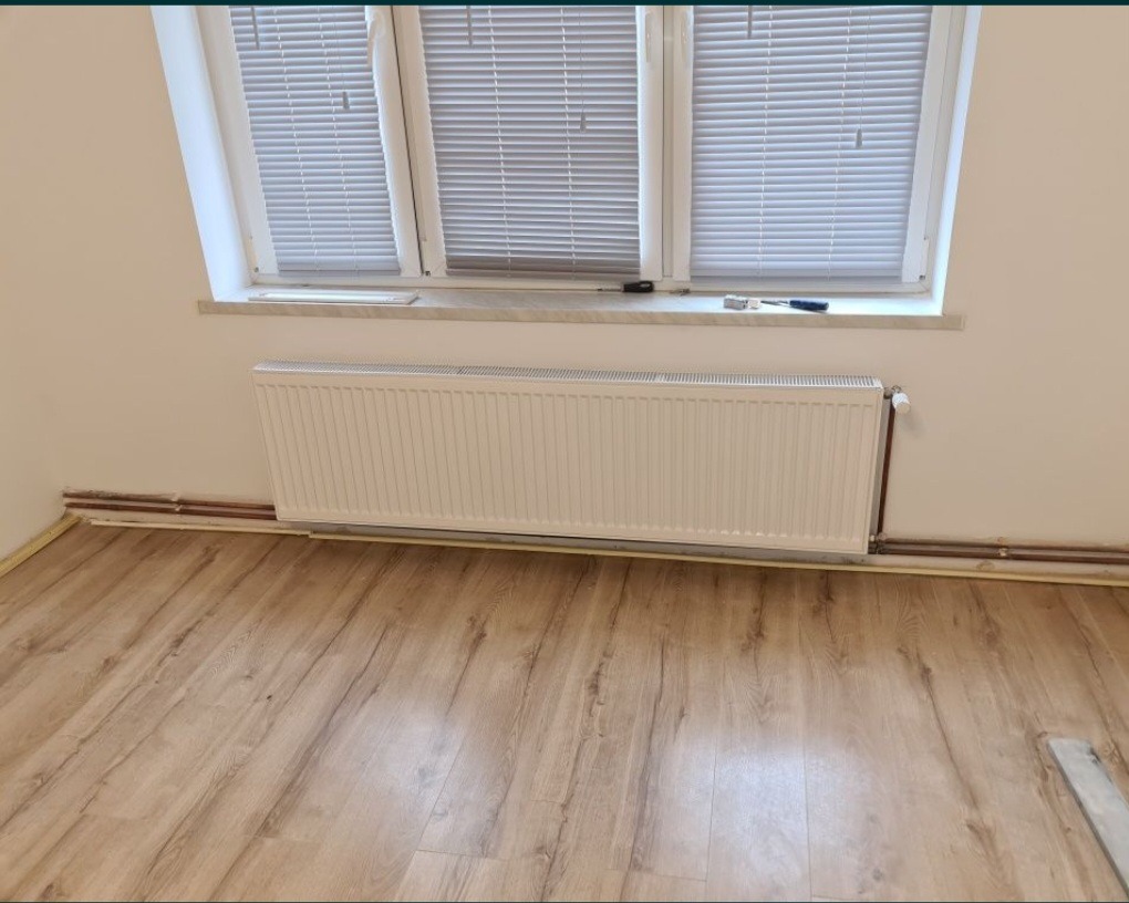 Nowy, biały grzejnik panelowy zamontowany przy ścianie pod oknem z żaluzjami, widoczne miedziane rury instalacji centralnego ogrzewania i fragment podłogi wyłożonej panelami imitującymi drewno.